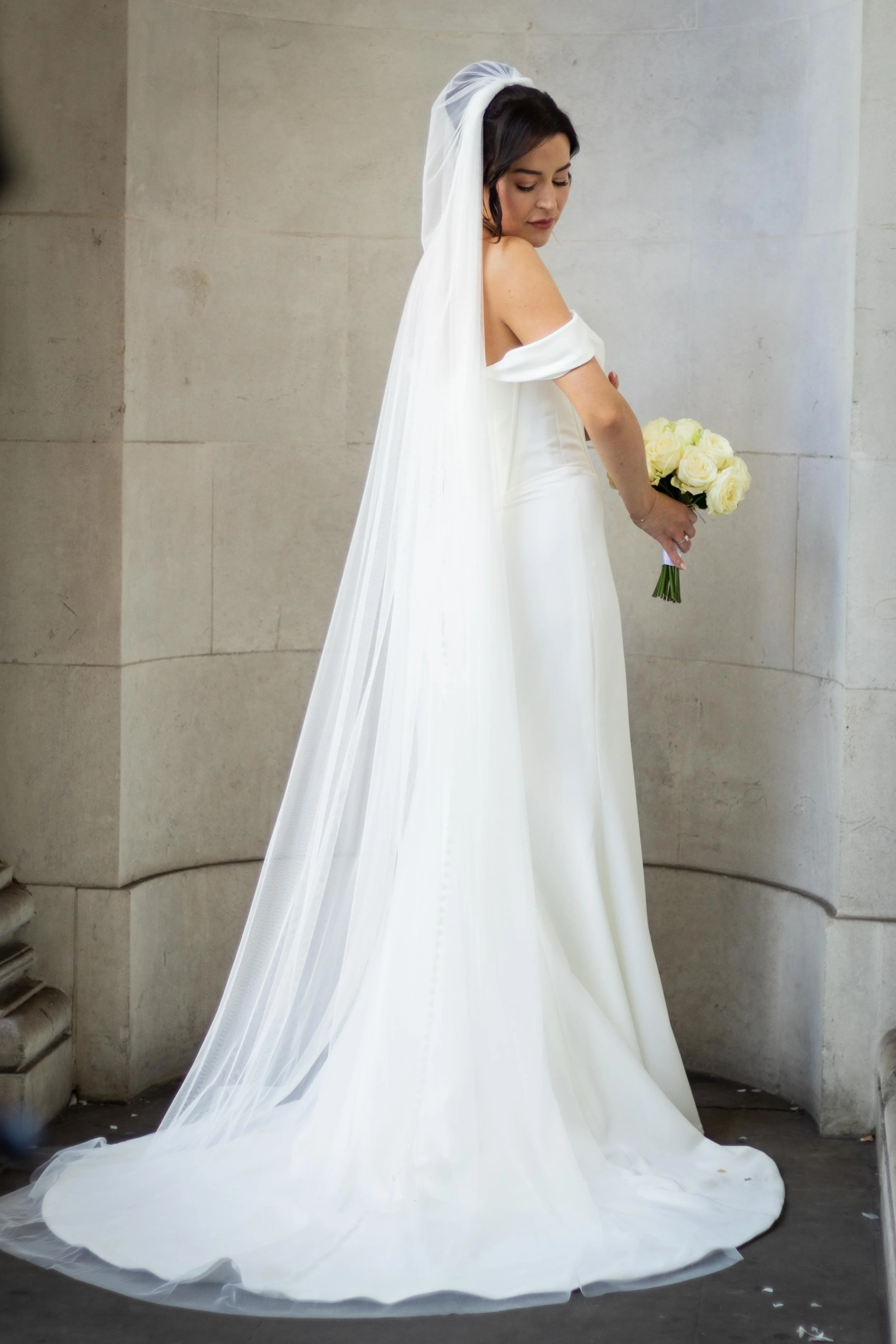 A bride standing against a stone wall, wearing a white wedding gown with a long veil, holding a bouquet of white roses, and looking down.