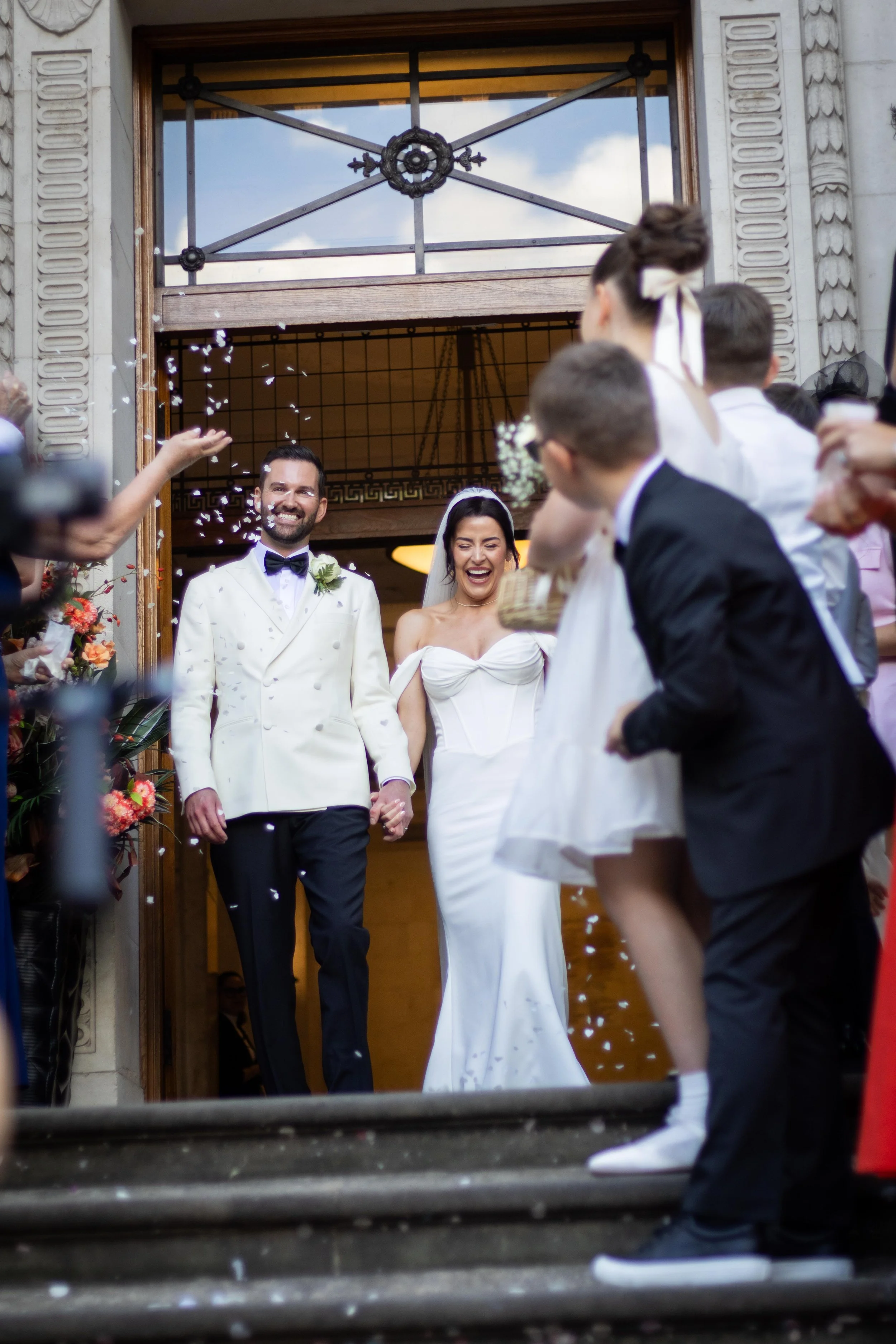 A newly married couple exiting a building, holding hands and smiling as guests celebrate with confetti. The groom wears a white tuxedo with a bow tie, and the bride wears a white wedding dress with a veil. Guests surround them, throwing confetti and 
