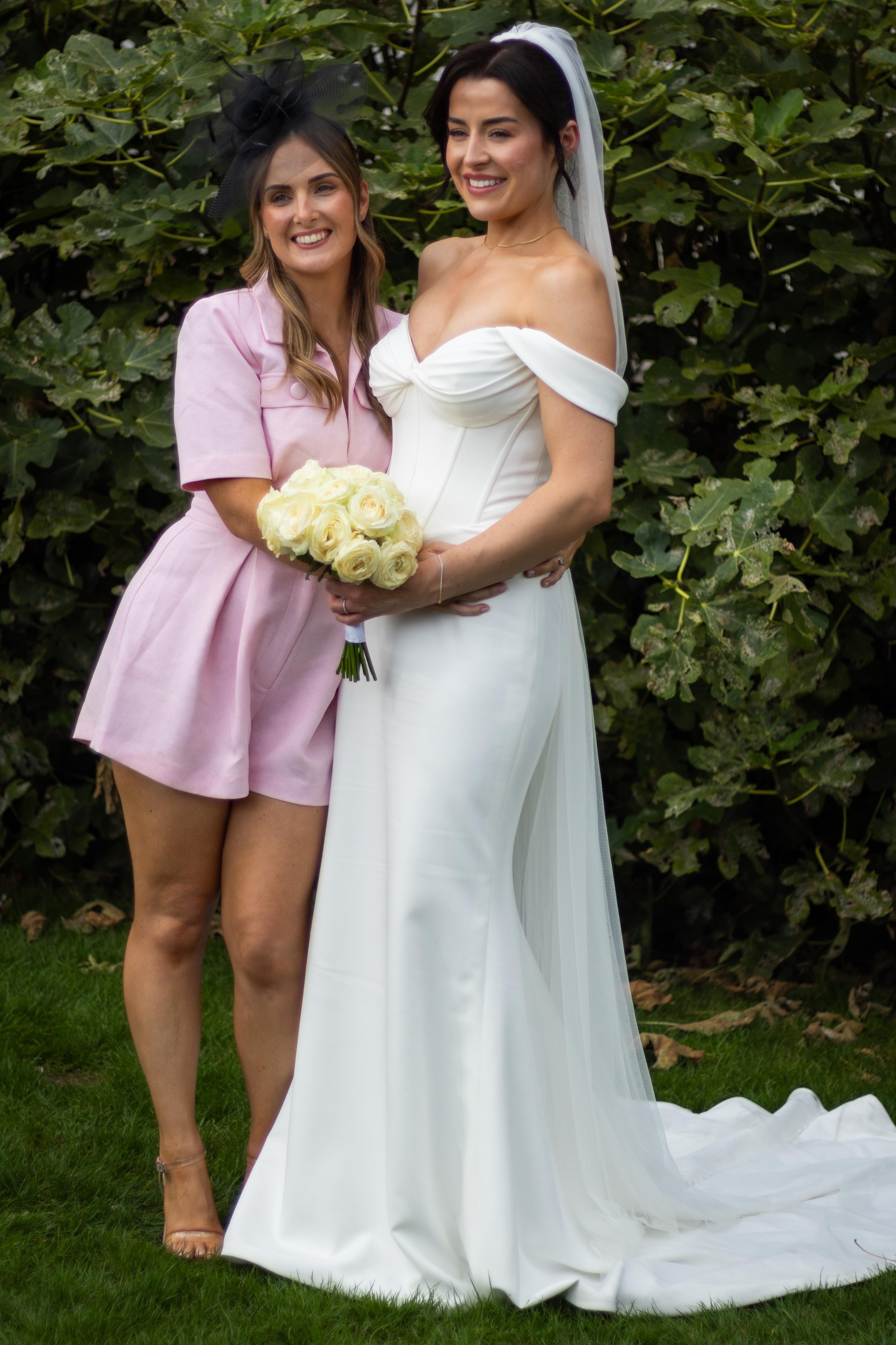 Two women, one in a white wedding gown and the other in a pink dress, standing outdoors in front of greenery. The woman in the wedding gown is holding a bouquet of white roses and wearing a veil. They are smiling and posing together, celebrating a we