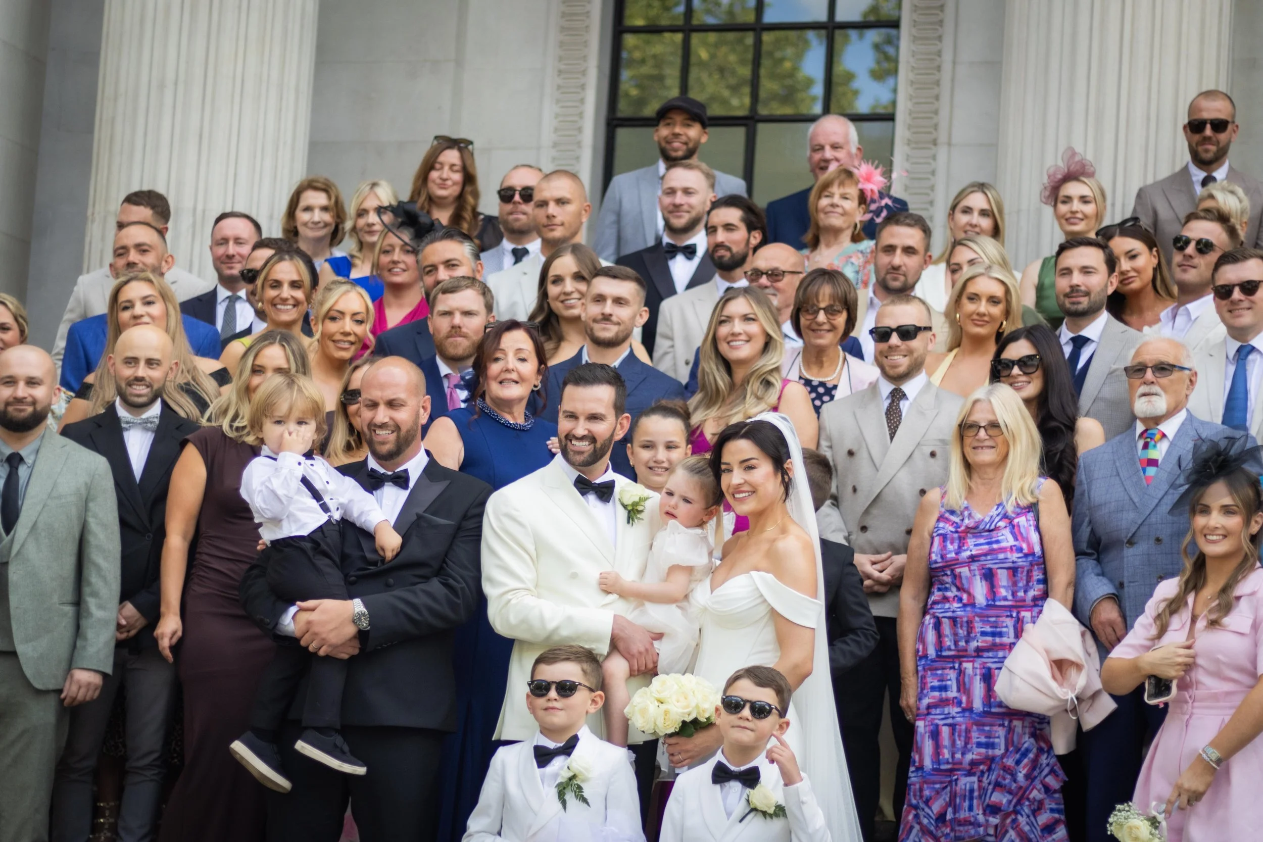 Large group of people in formal attire posing for a photo on a building steps, likely at a wedding celebration.