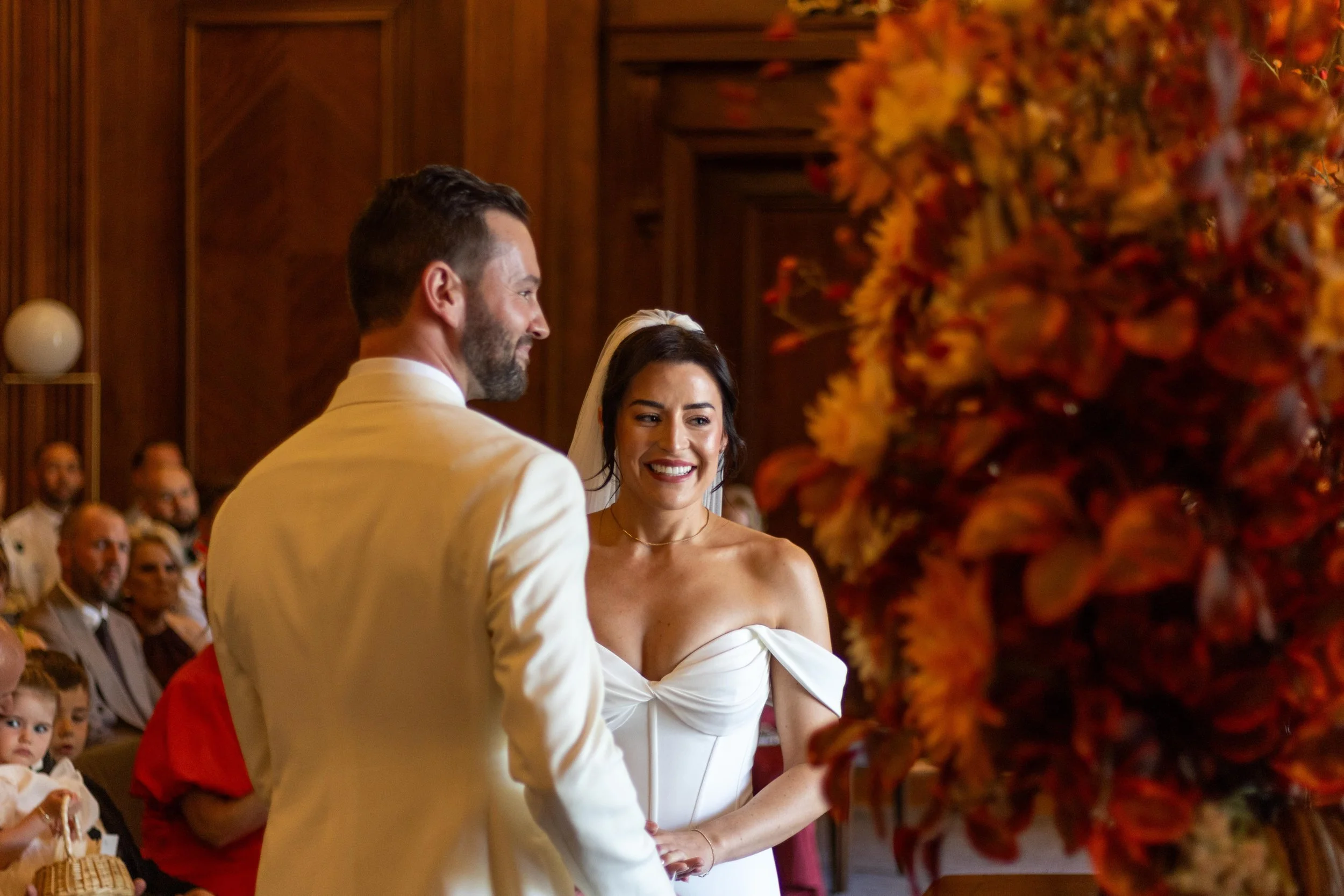 A bride and groom exchanging vows during their wedding ceremony in a church or chapel with wooden walls, surrounded by guests. The bride is smiling and wearing a strapless white wedding dress, while the groom is dressed in a white suit.