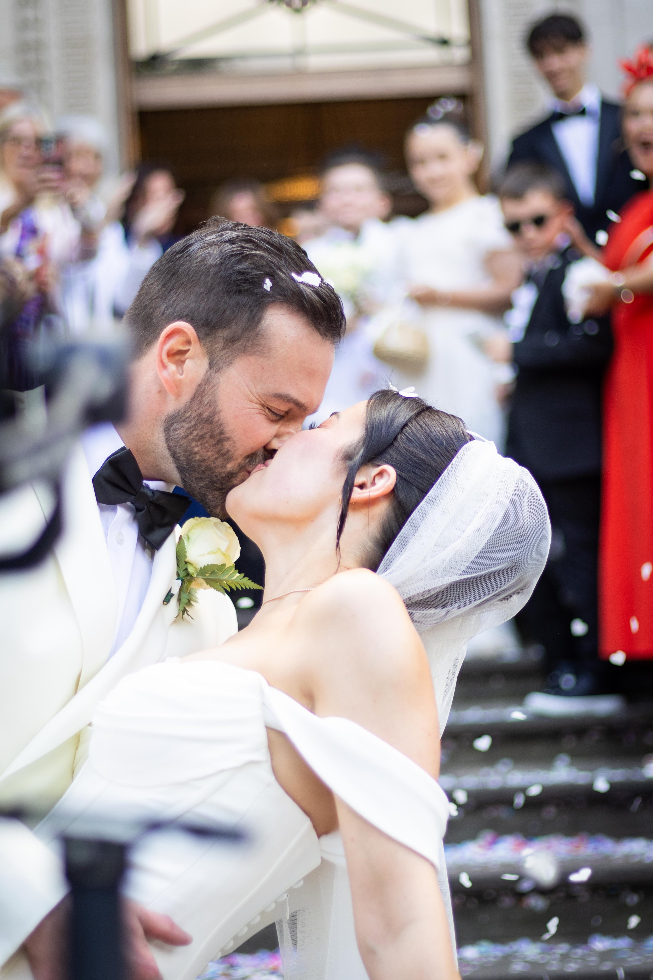 A newlywed couple shares a kiss during their wedding ceremony outside with friends and family in the background, surrounded by falling flower petals.