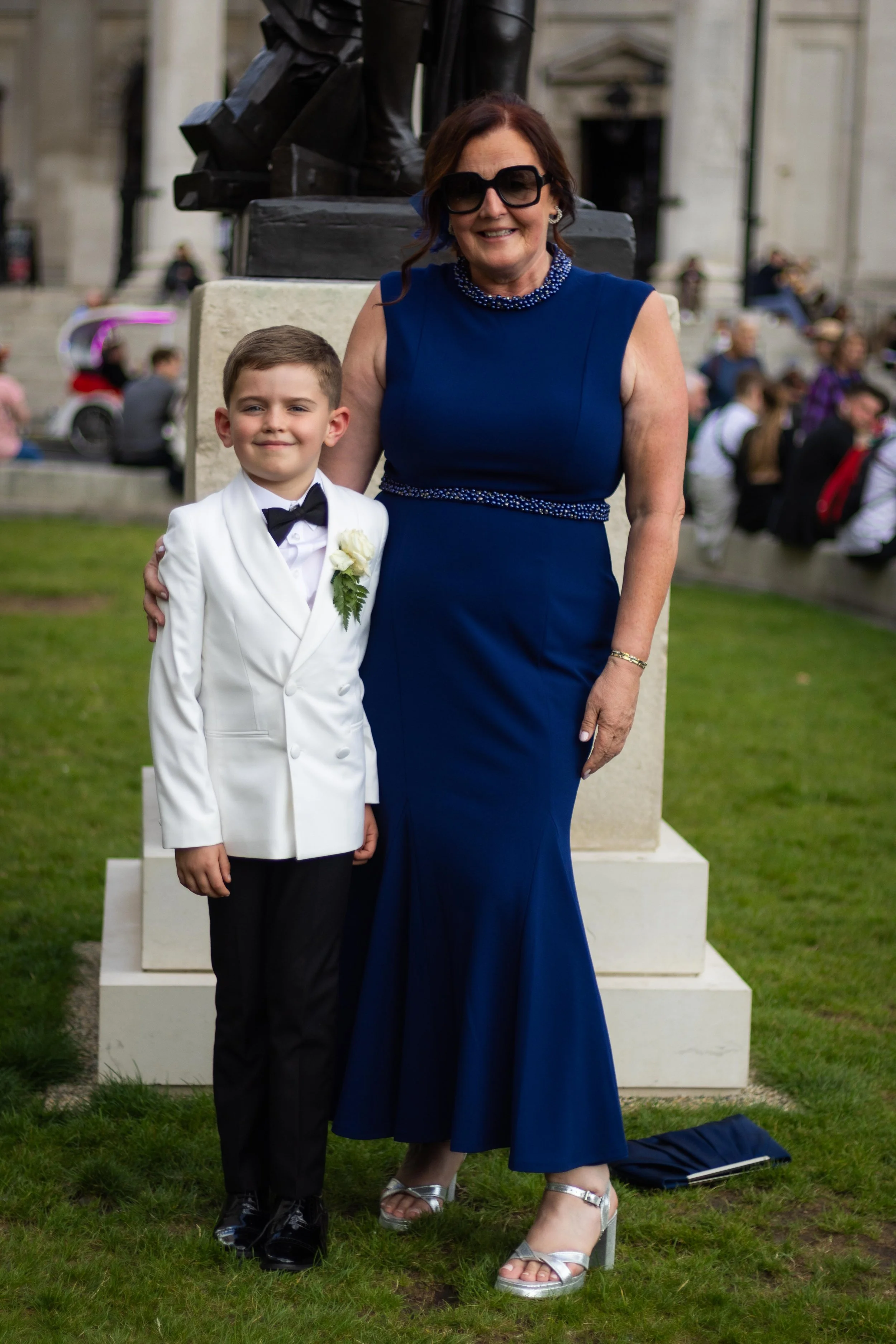 A woman in a blue dress and black sunglasses standing next to a young boy in a white tuxedo at a formal outdoor event.