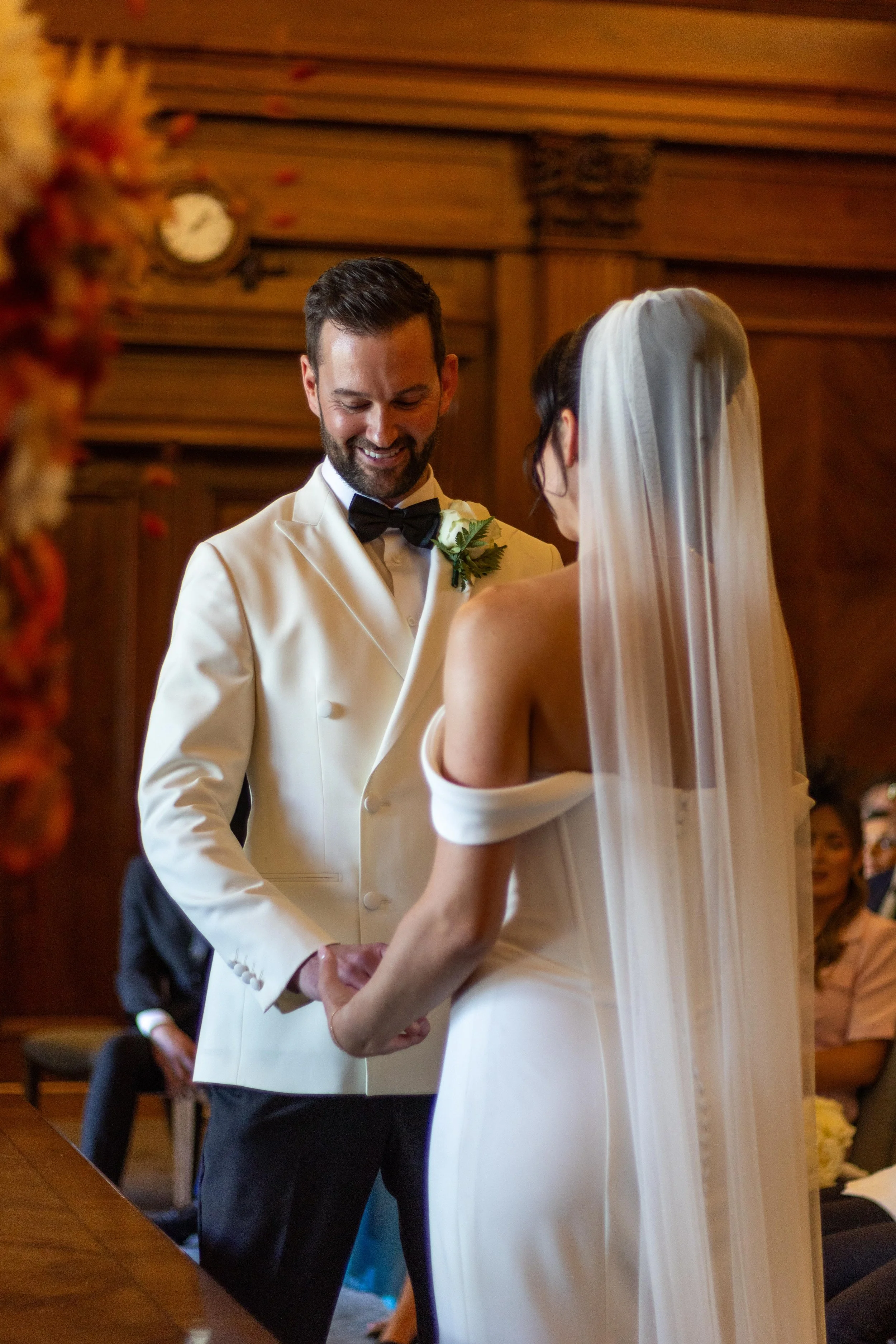 A groom and bride holding hands during a wedding ceremony, with the groom smiling and the bride facing away, wearing a wedding dress and veil inside a wood-paneled room.