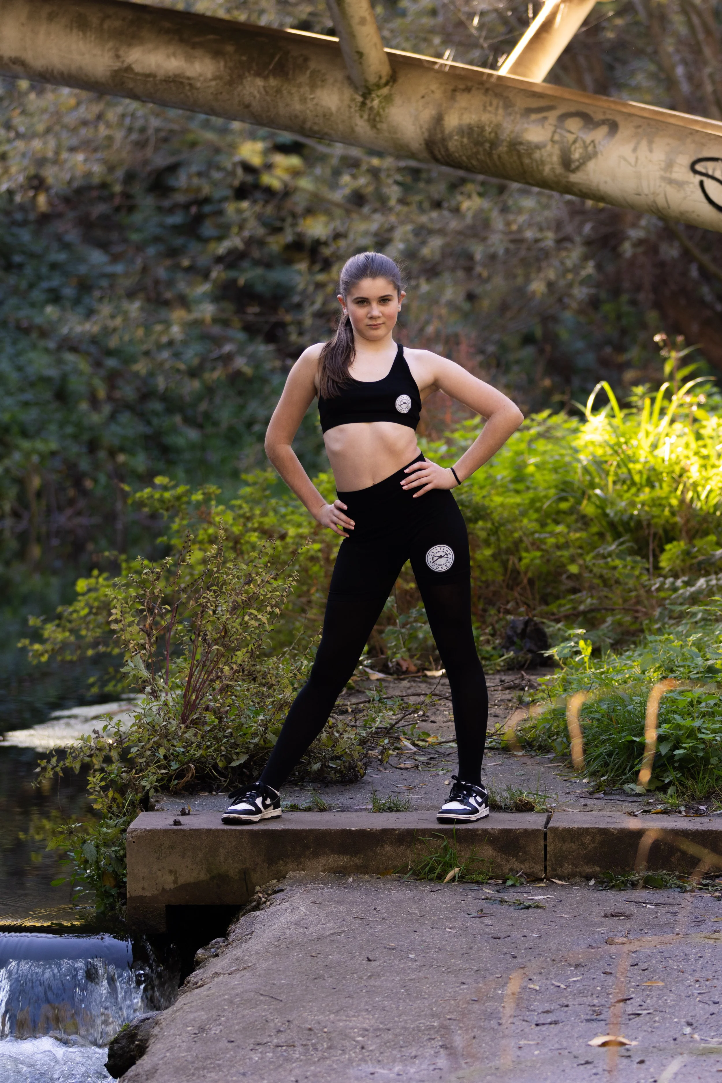 A young woman in black athletic wear, standing on a concrete ledge near water, outdoors with green foliage in the background.