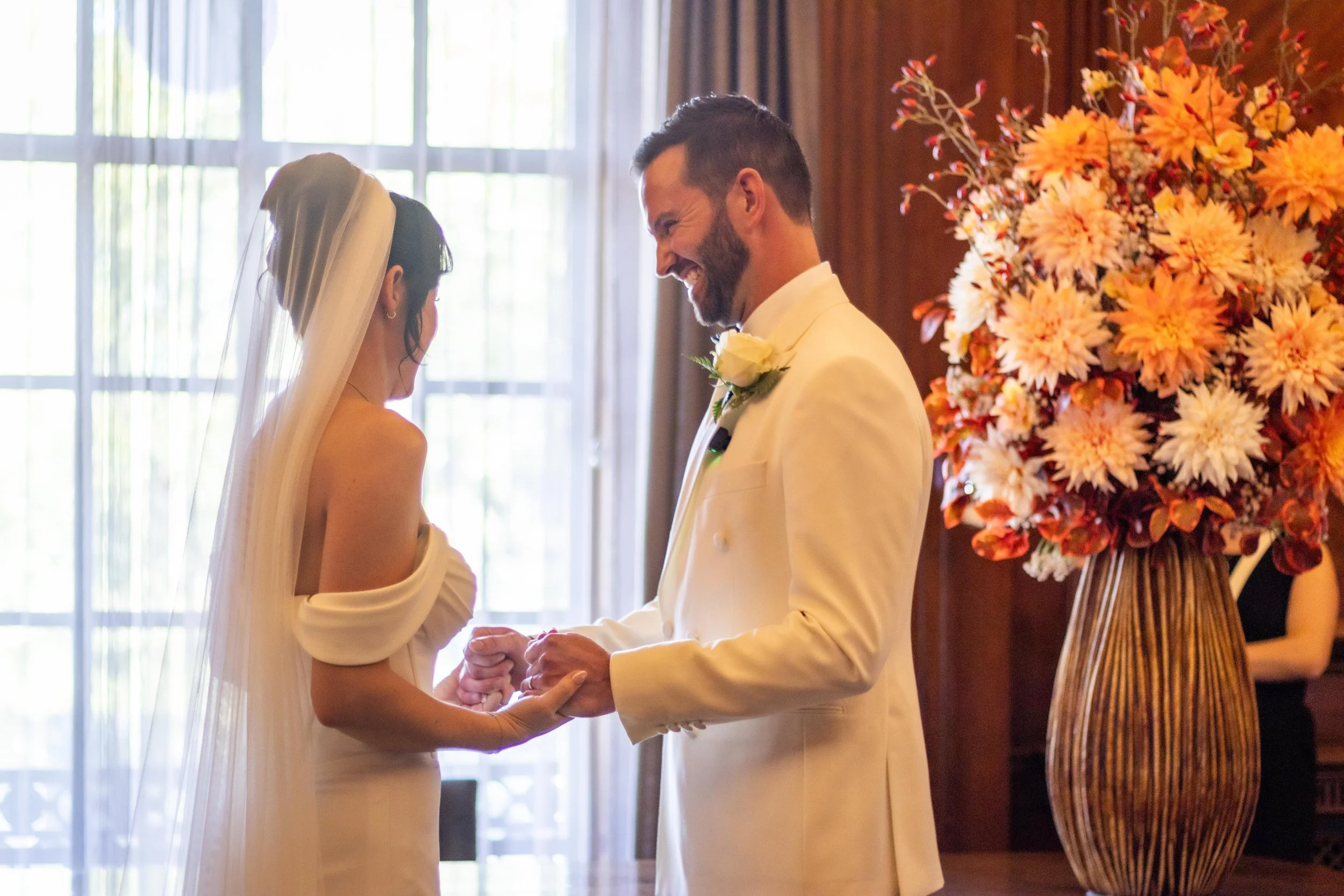 A bride and groom during their wedding ceremony exchanging vows and holding hands inside a decorated room with large windows and a flower arrangement.