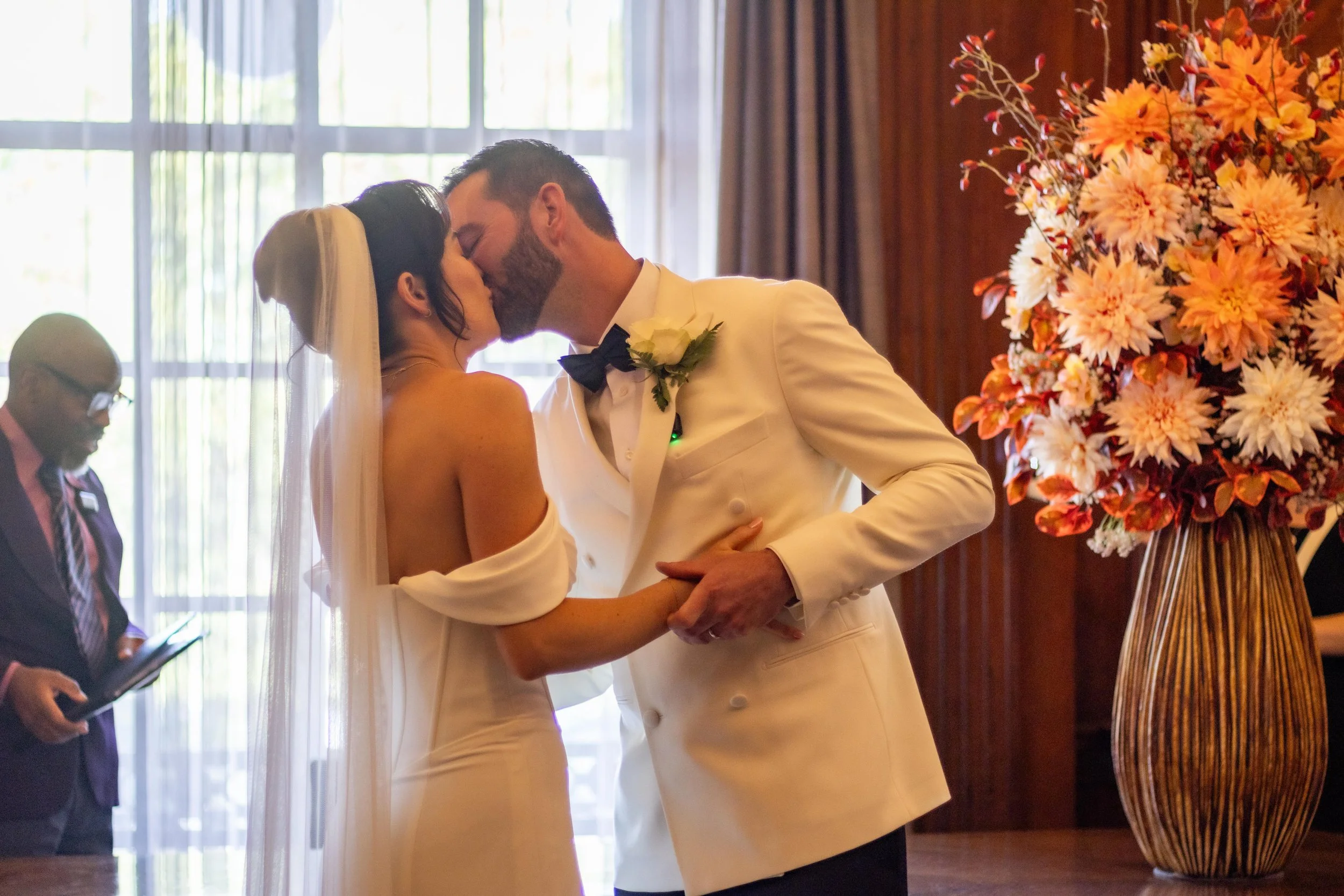 A bride and groom kiss during their wedding ceremony indoors, with a woman in the background reading from a tablet, and a large floral arrangement nearby.