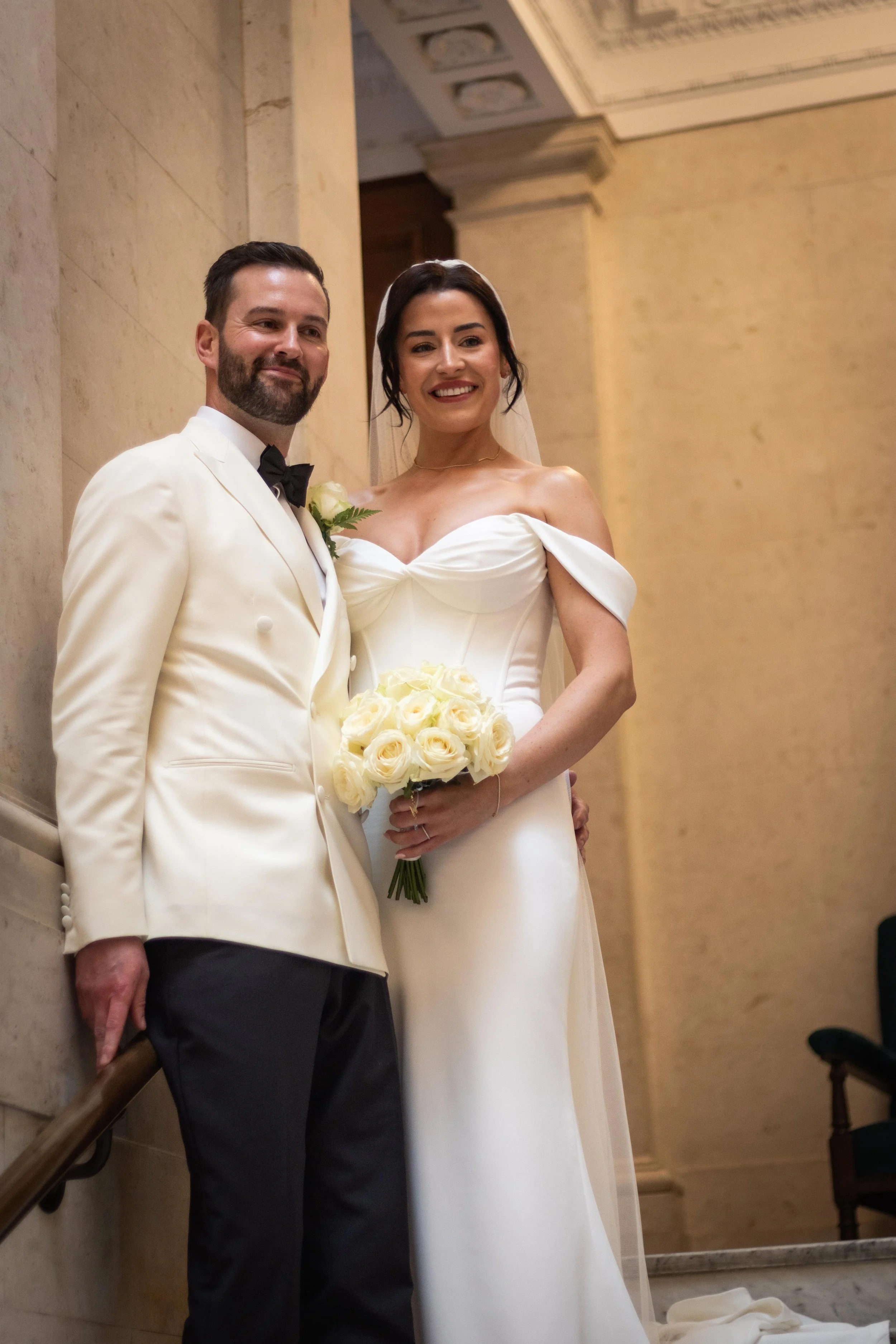 A bride and groom standing together at their wedding in an elegant indoor setting, with the bride holding a bouquet of cream roses.