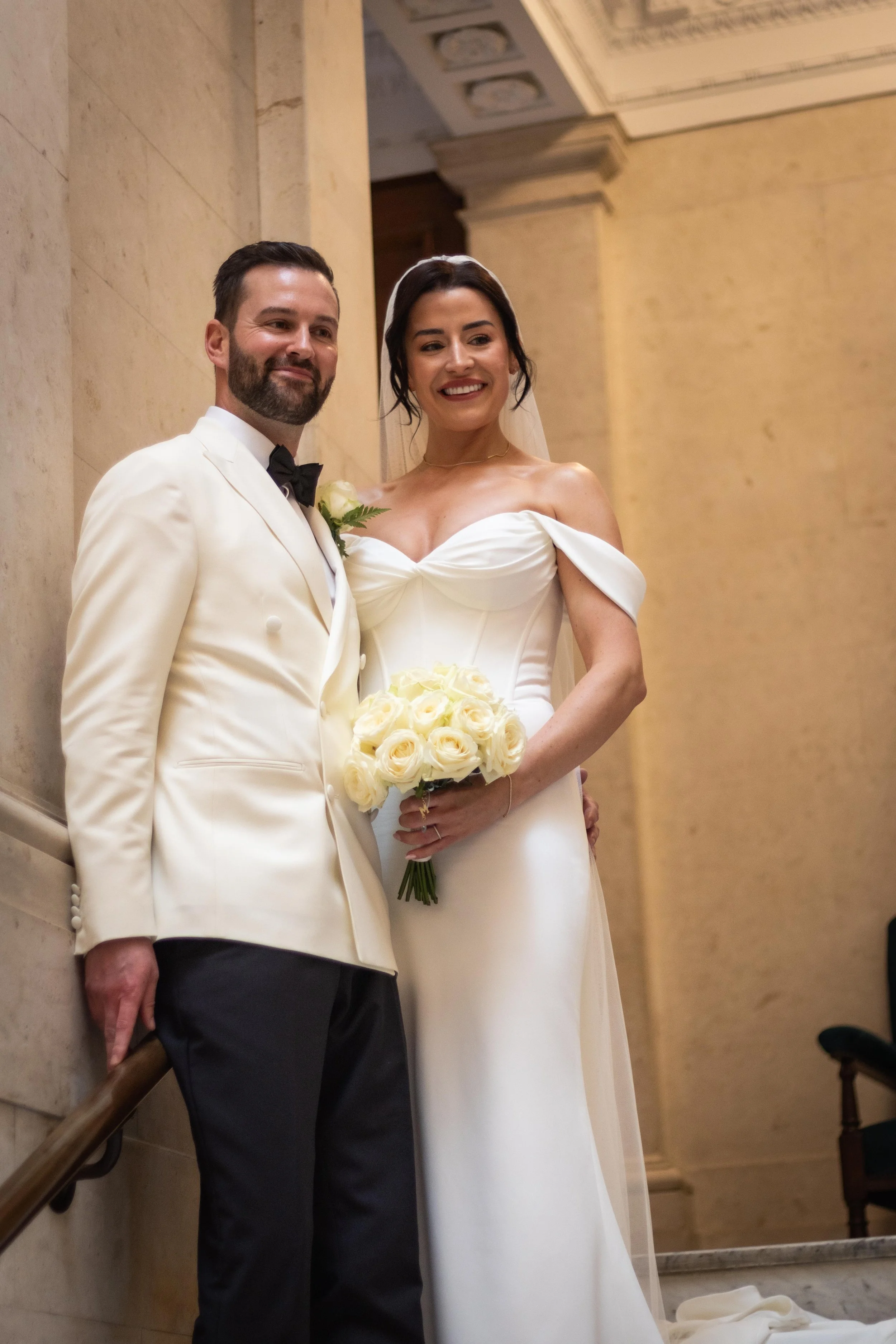 A bride and groom in wedding attire standing together indoors, with the bride holding a bouquet of white roses.