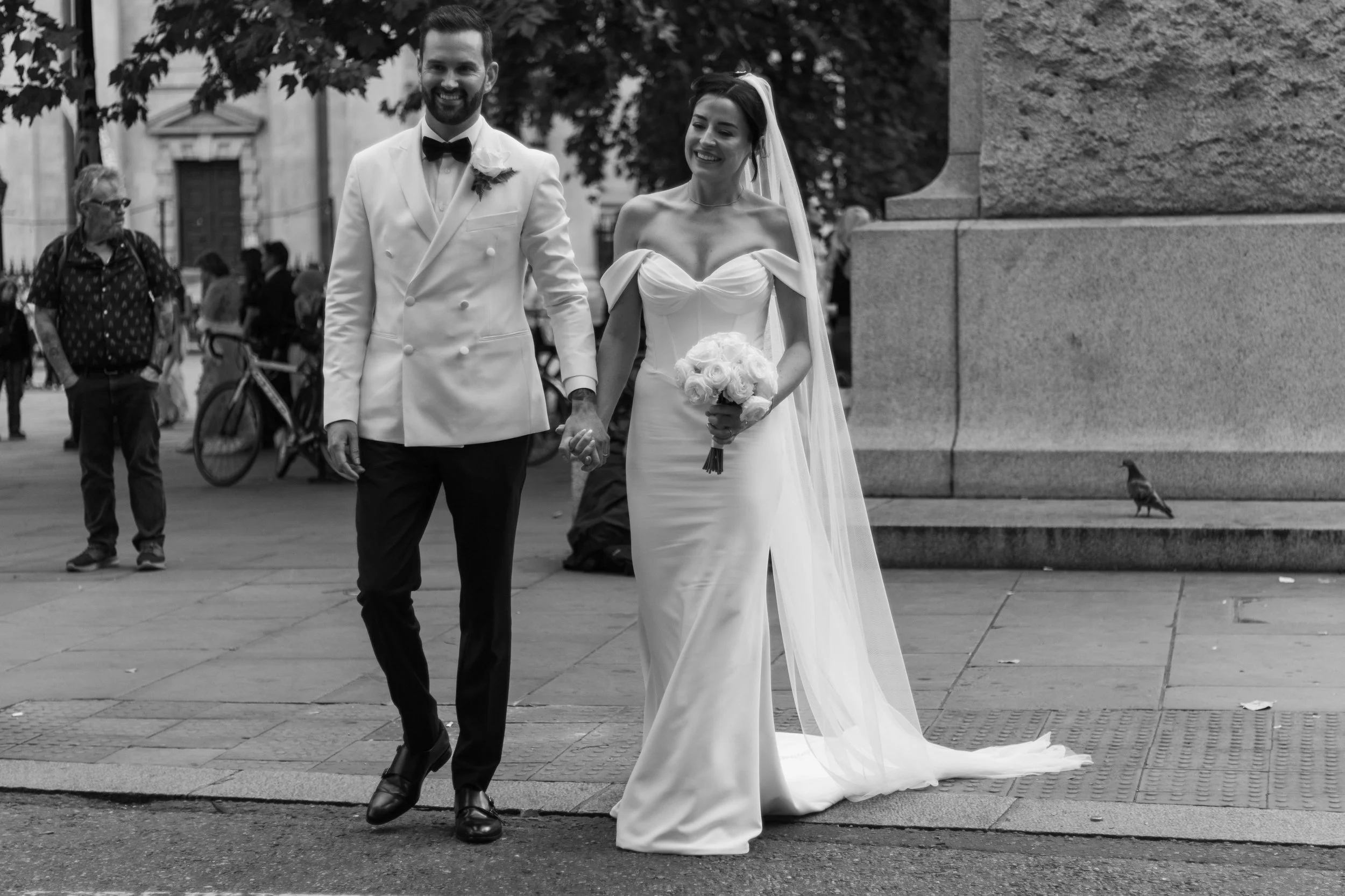 A bride and groom holding hands and walking outdoors on a city street, dressed in wedding attire, smiling, with pedestrians and a pigeon in the background.