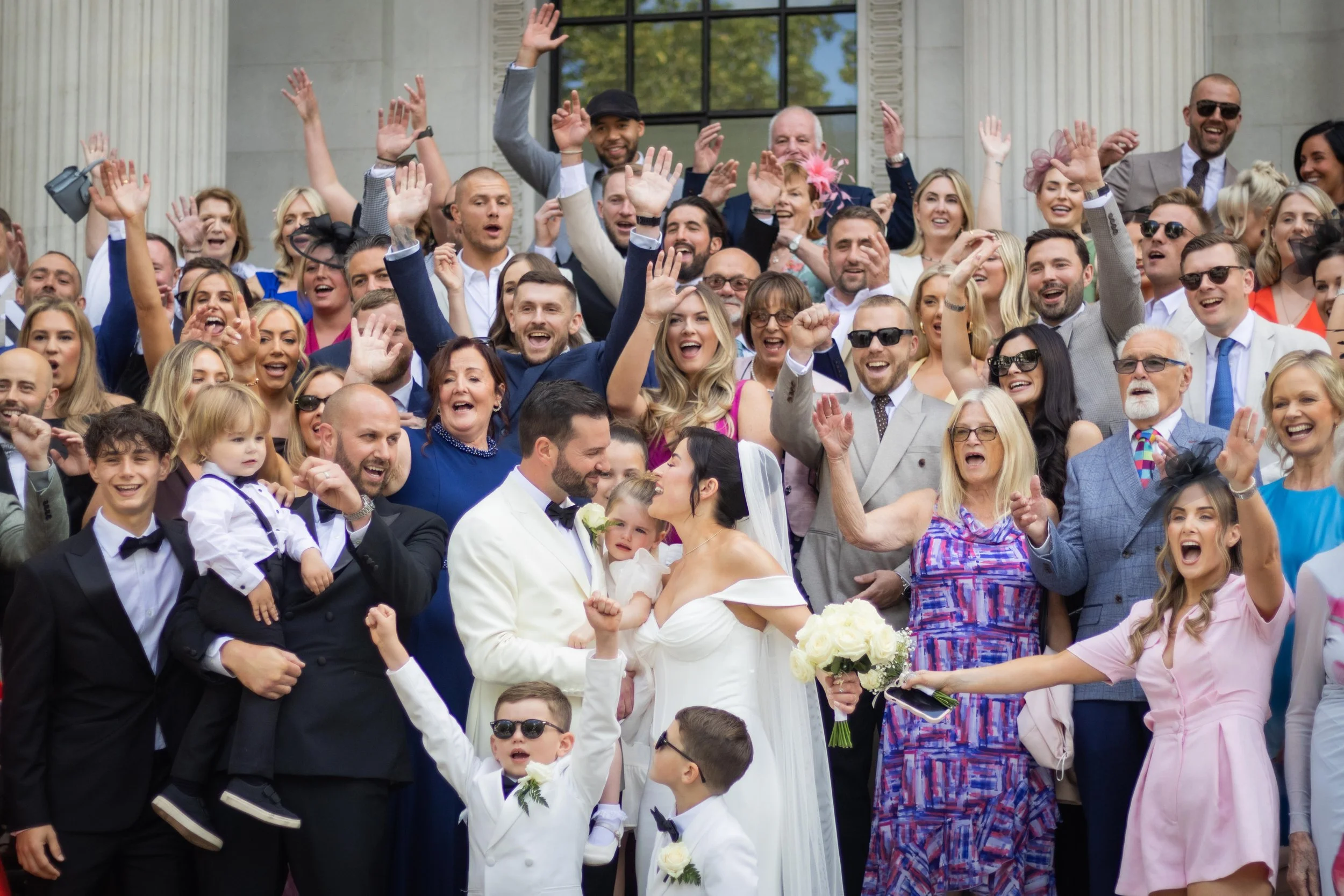 A large group of people celebrating a wedding outdoors. The bride and groom are in the center, surrounded by family and friends, all smiling, cheering, and raising their hands.