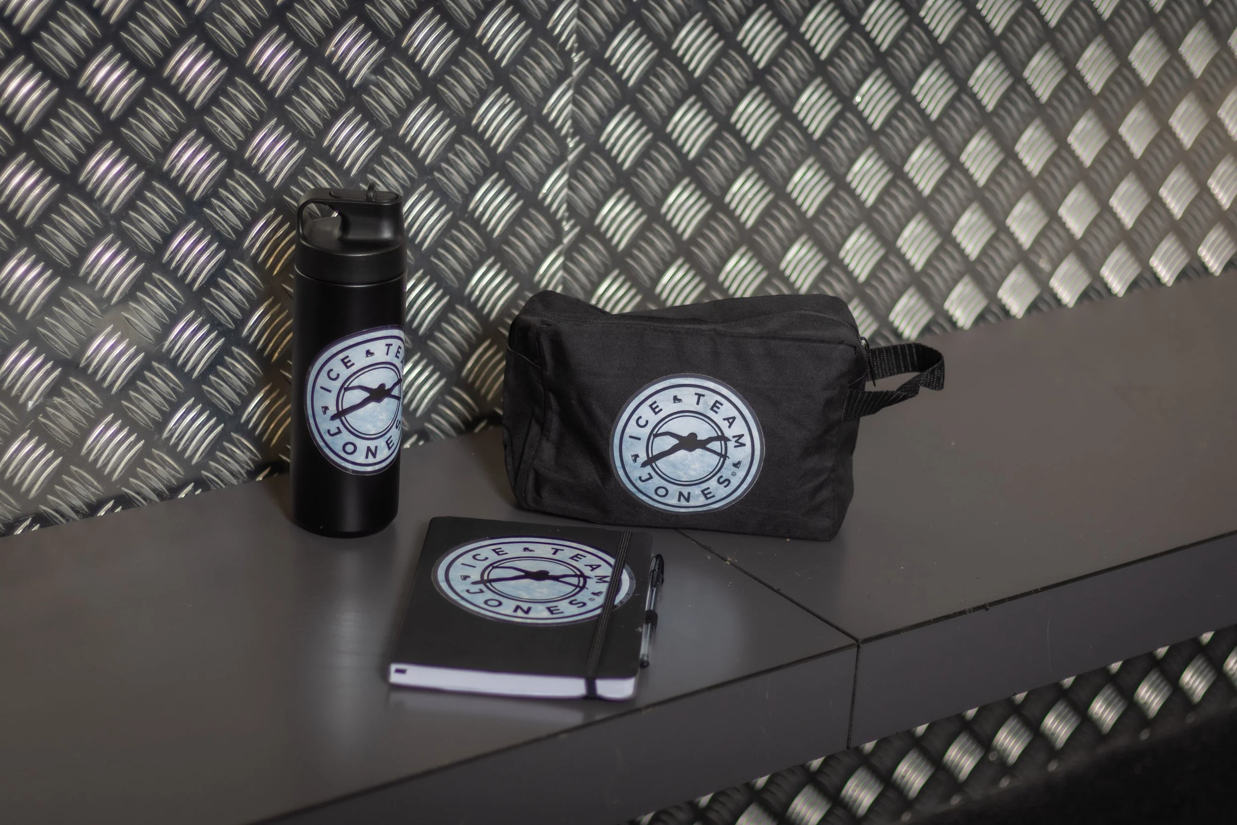Black travel bag, water bottle, notebook, and pen on a grey table against a metallic textured wall, all with Team Ice logo.