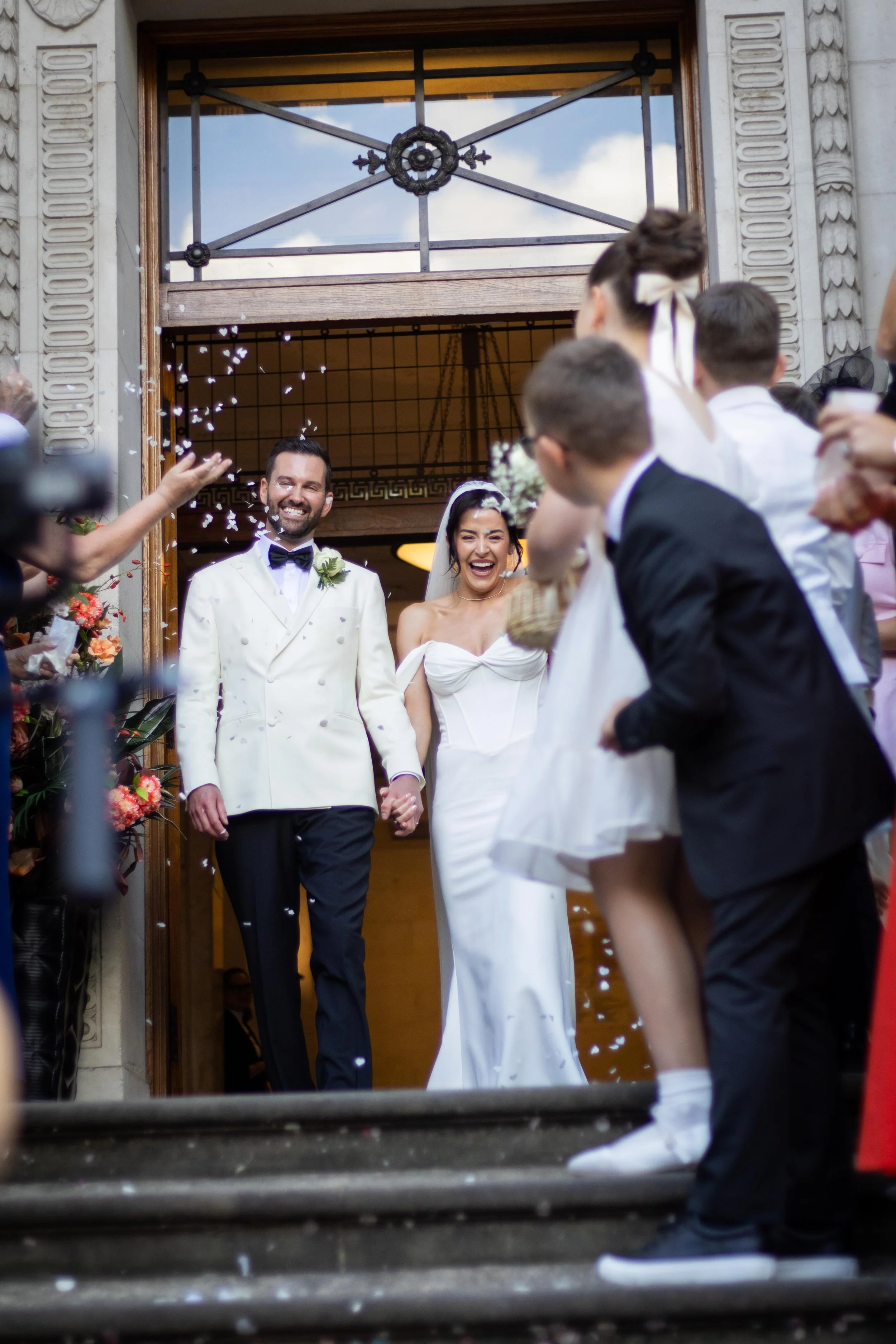 A newlywed couple exiting a building, holding hands and smiling, surrounded by friends and family celebrating with confetti.