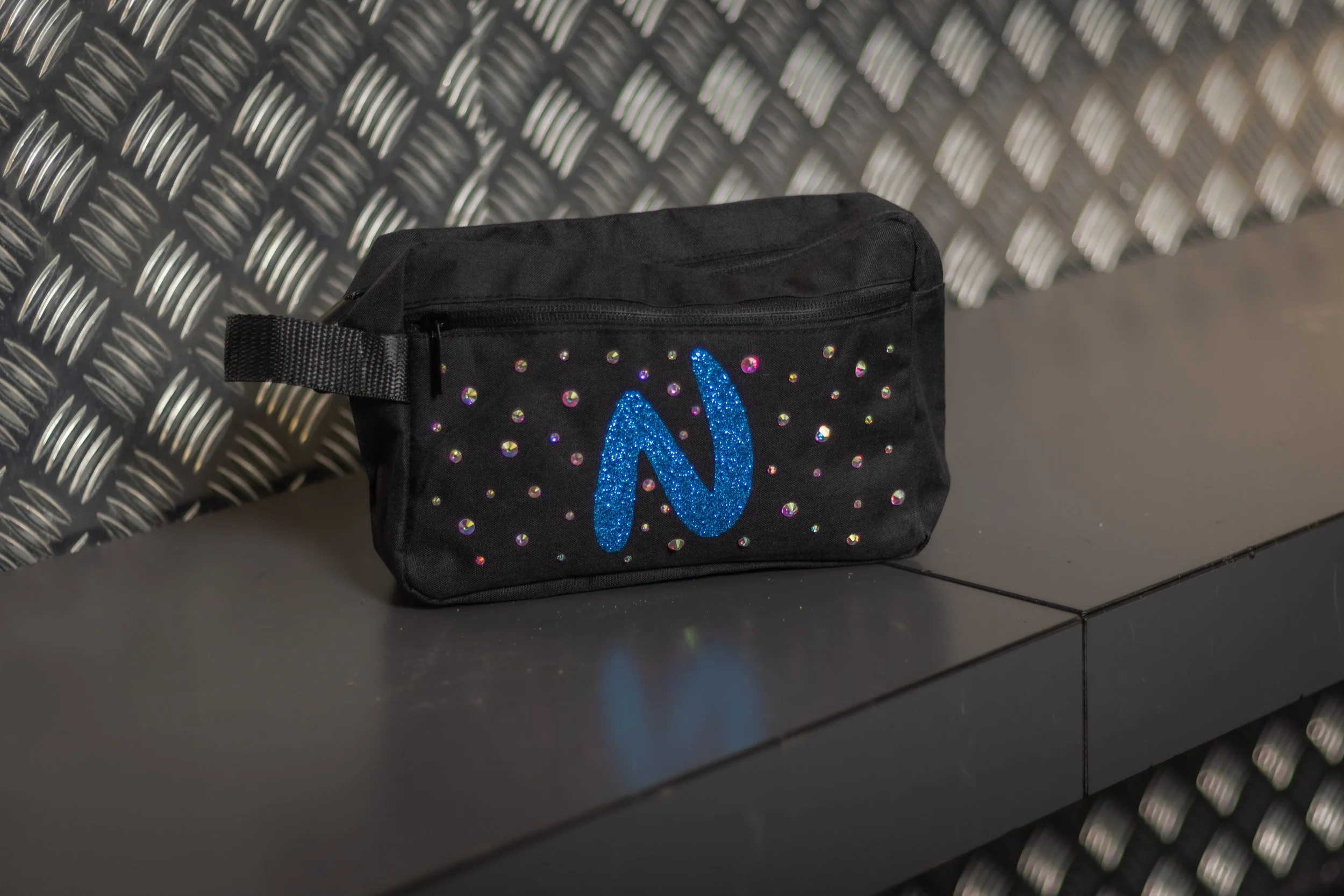 A black small pouch with a large blue glitter letter 'N' on the front, decorated with small iridescent rhinestones, resting on a dark gray surface against a textured metal wall.