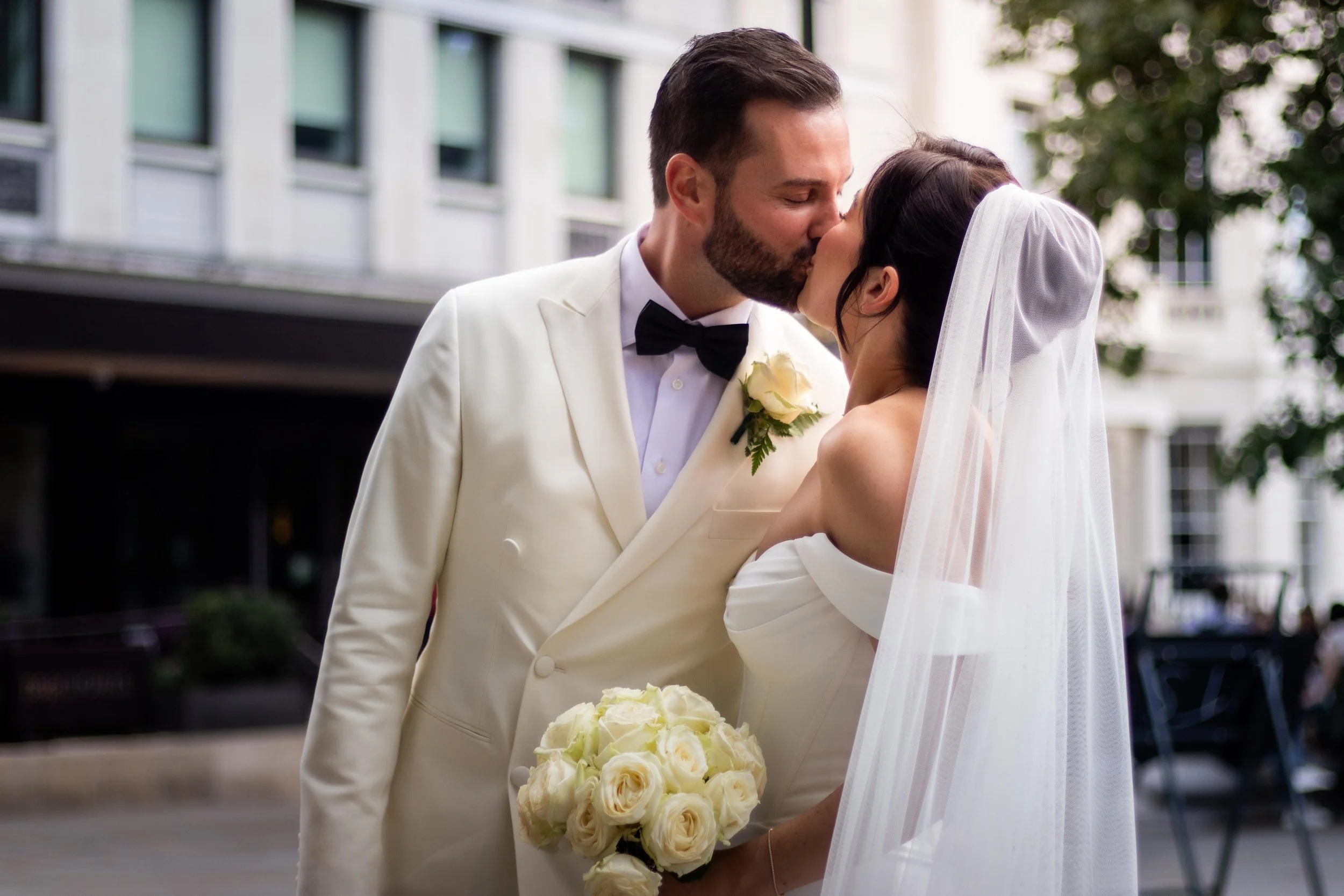 A newlywed couple sharing a kiss outdoors in a city, the bride holding a bouquet of white roses, the groom wearing a tuxedo with a boutonnière, in front of modern buildings and trees.