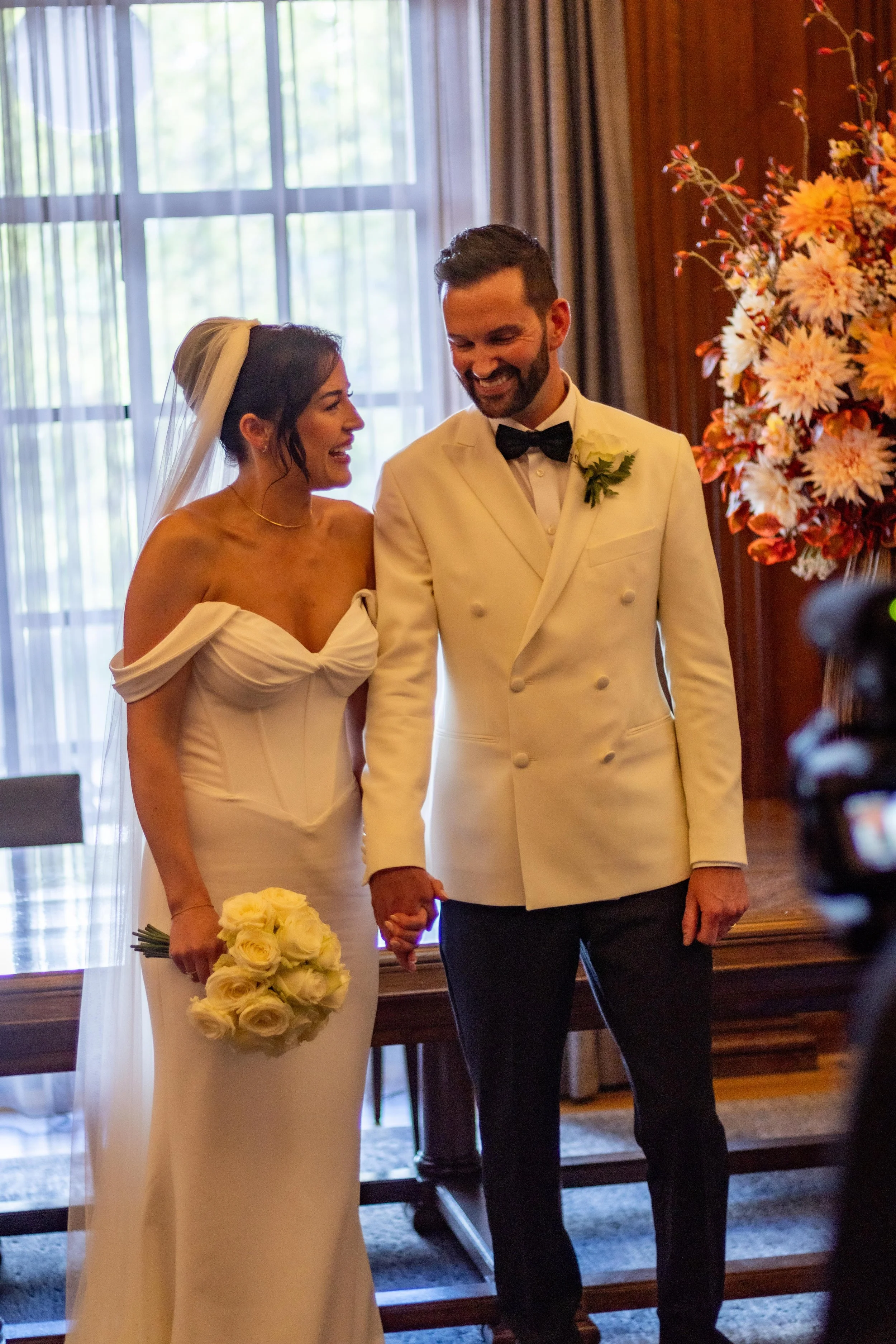 Bride and groom standing together during their wedding ceremony, holding hands, in front of a window with curtains. The bride is wearing a white off-shoulder wedding dress and holding a bouquet of white roses. The groom is wearing a white tuxedo jack