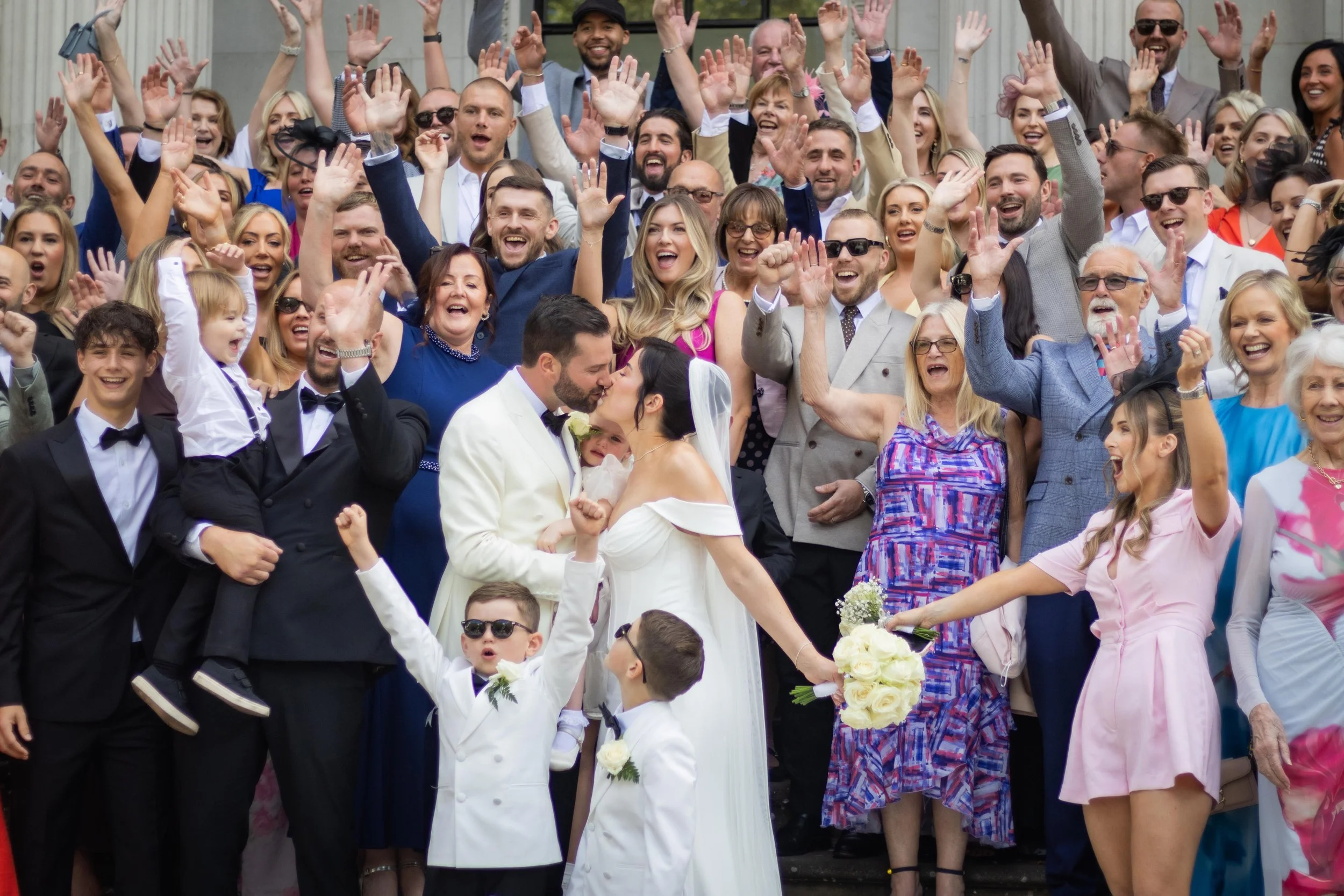 A large group of people gathered outside, celebrating a wedding, with many raising their hands and smiling; the bride and groom are kissing in the center.