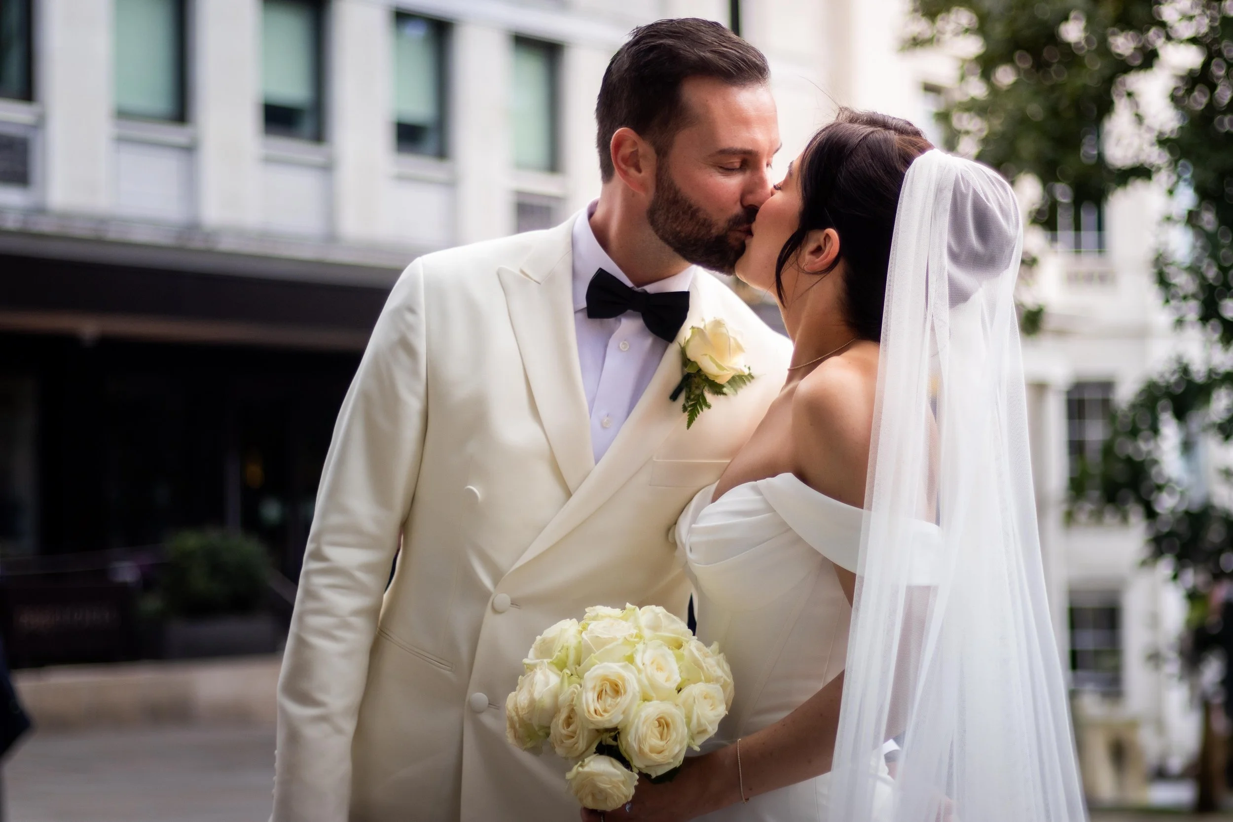 A bride and groom kissing outdoors, with the bride holding a bouquet of white roses, and the groom wearing a cream tuxedo with a black bow tie and a white rose boutonniere.