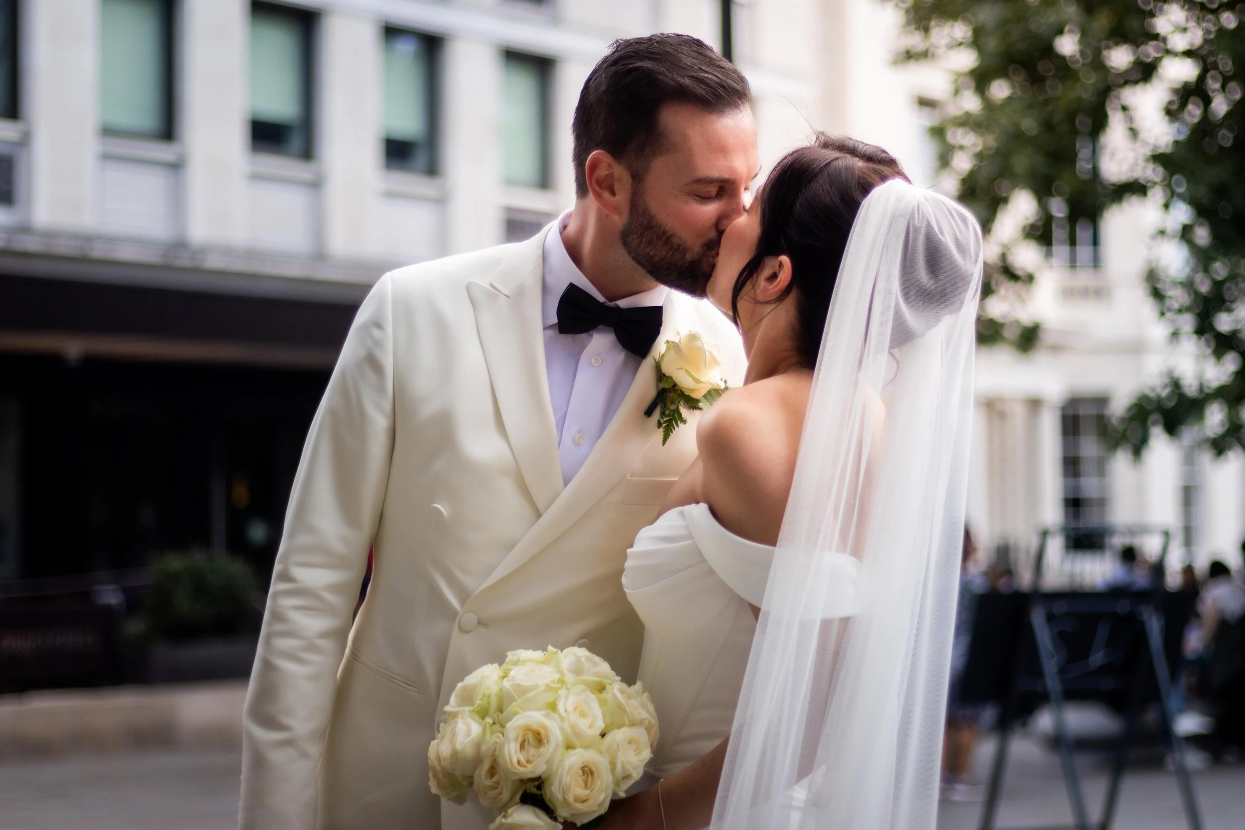A newlywed couple sharing a kiss outdoors, dressed in wedding attire, with the groom in a white tuxedo and the bride in a white off-the-shoulder gown holding a bouquet of white roses.