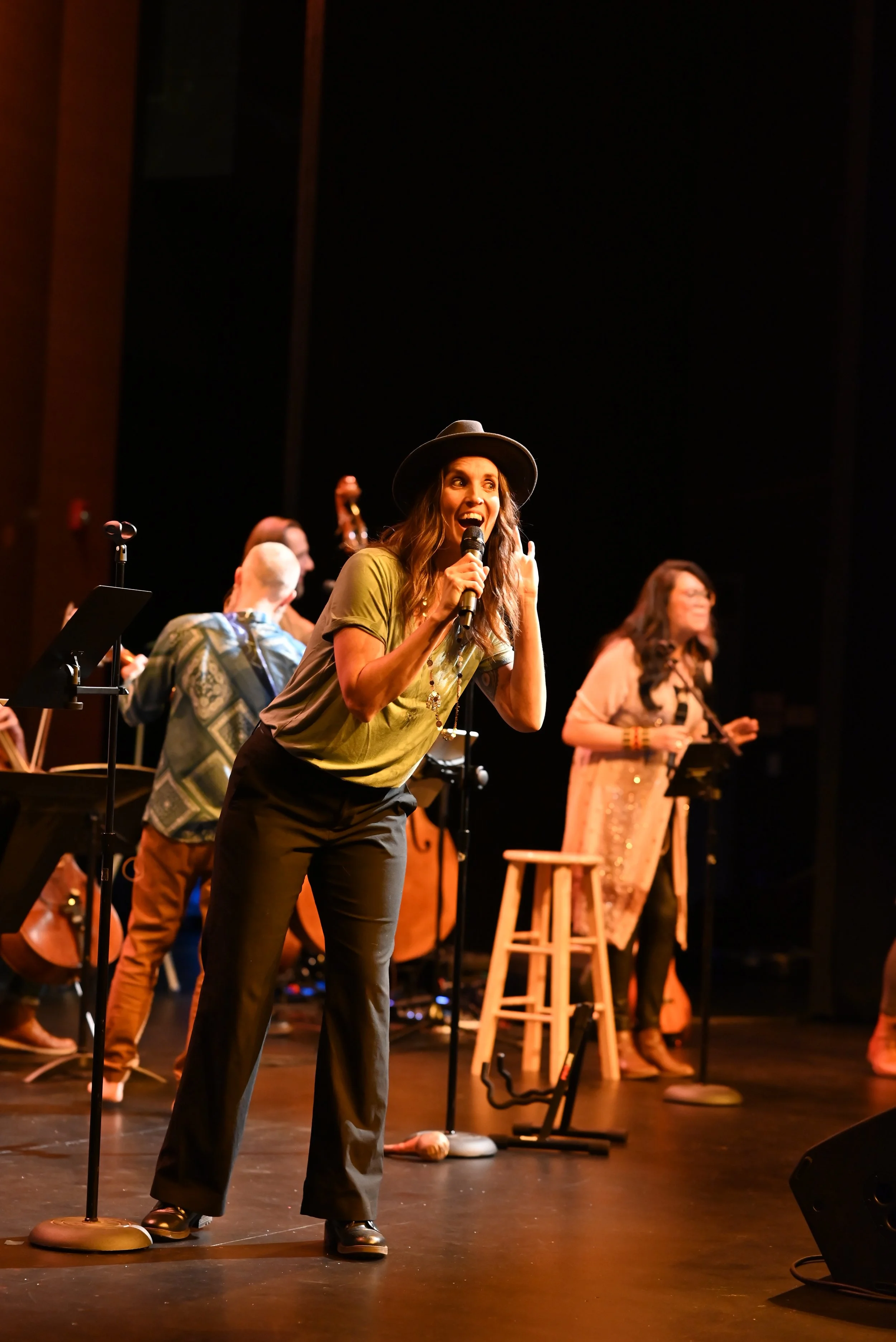 A woman wearing a black hat and green shirt performs on stage with a microphone, with backup singers and musicians in the background.