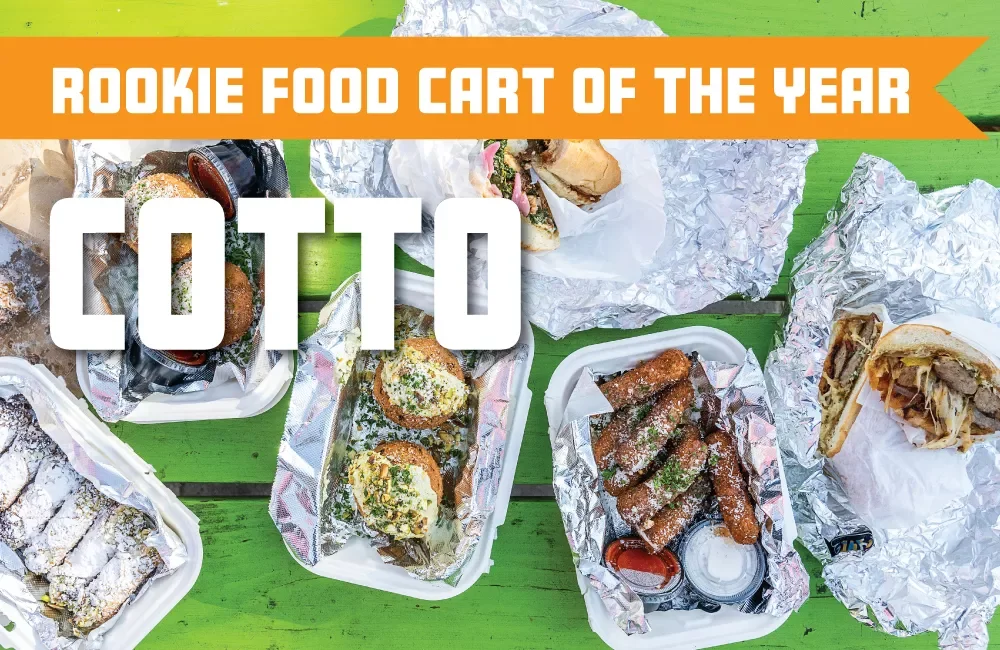 Assorted takeout food items including sandwiches, fried appetizers, and snacks on aluminum foil and styrofoam trays on a green wooden table.
