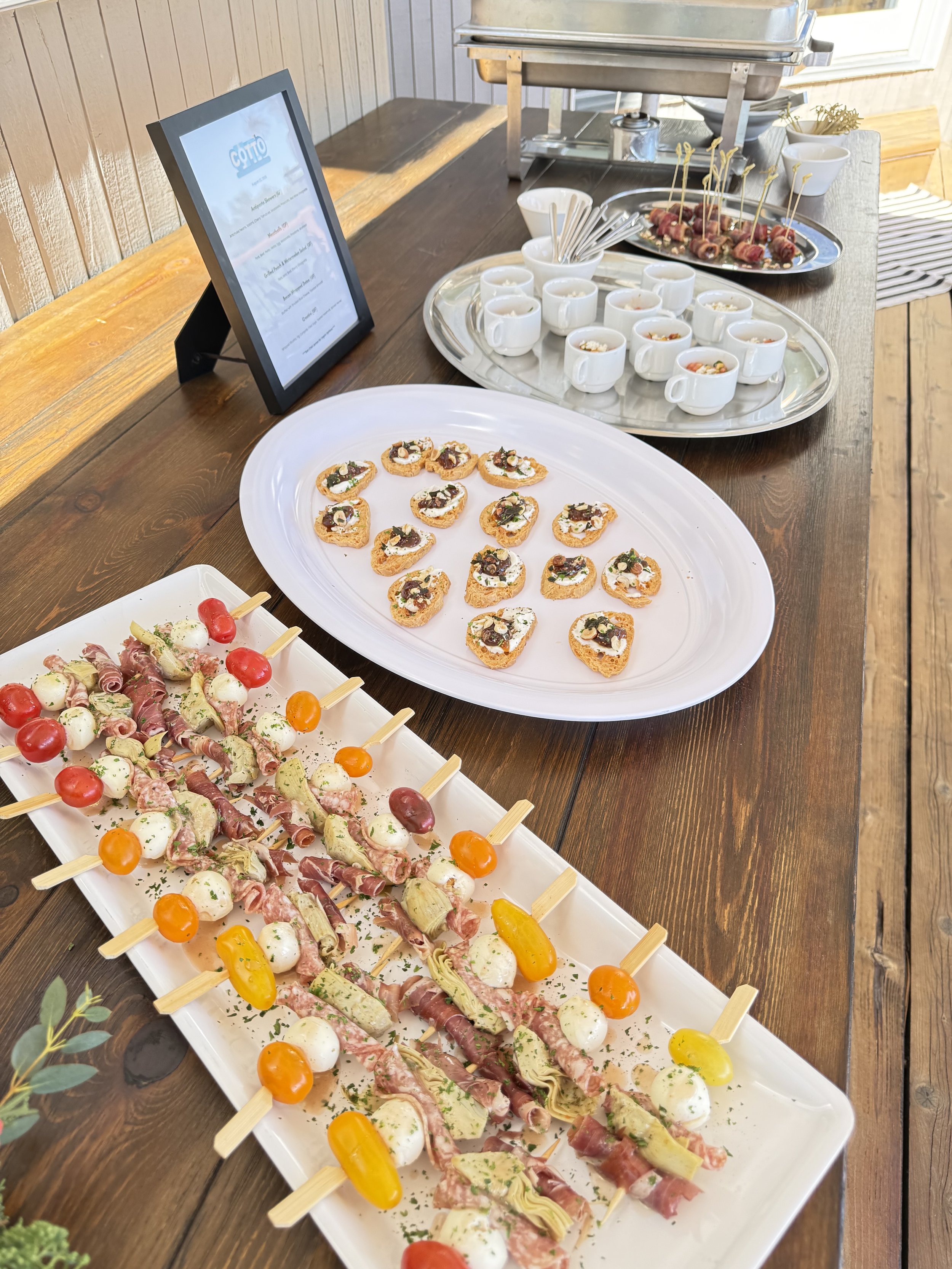 A buffet table with various appetizers including skewers of cherry tomatoes, cheese balls, sliced meats, and vegetables, along with small cups of soup or dip, and a tray of meat wrapped with toothpicks, set on a rustic wooden table.