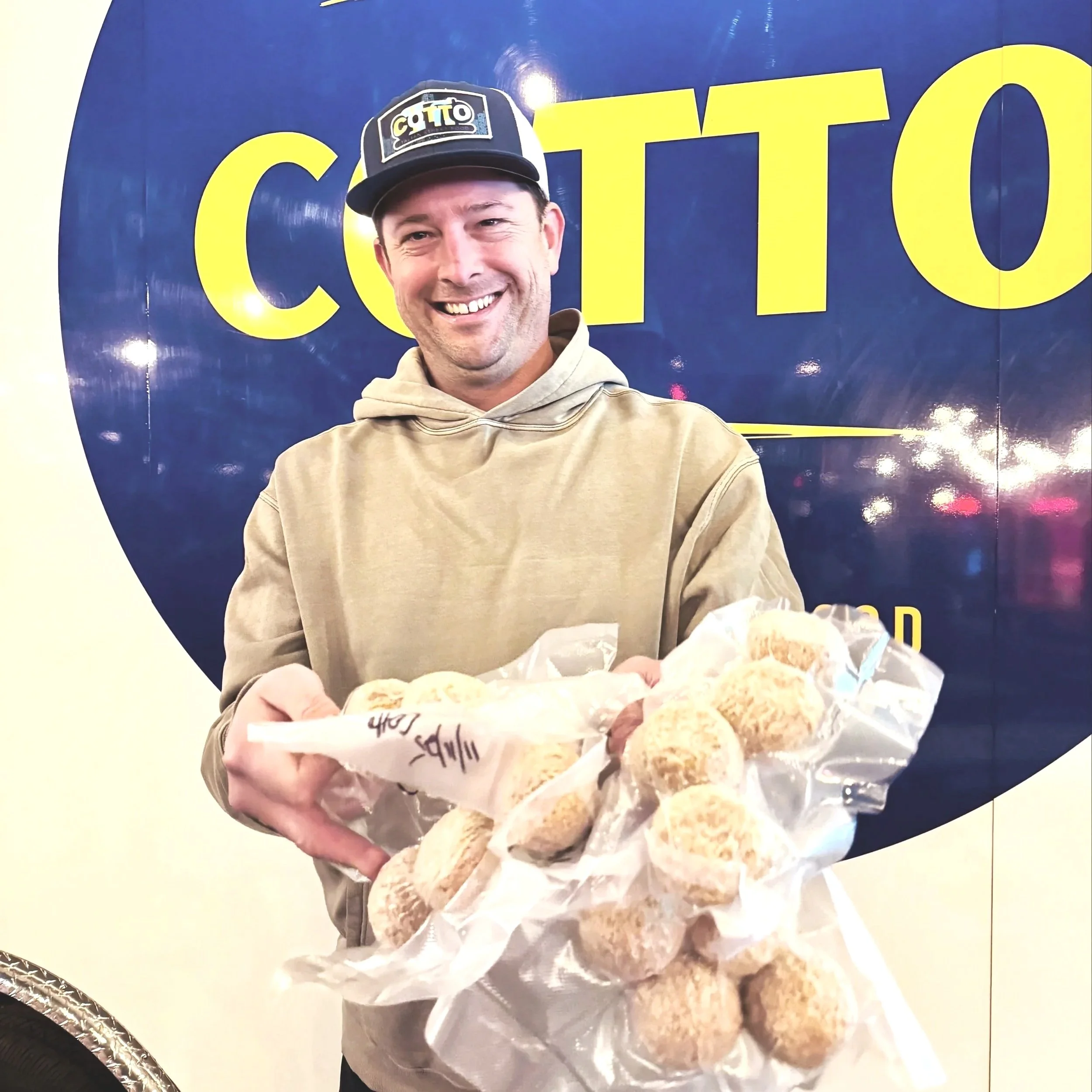 A smiling man holding a clear plastic bag filled with round, beige-colored objects, standing in front of a dark blue background with a yellow logo.