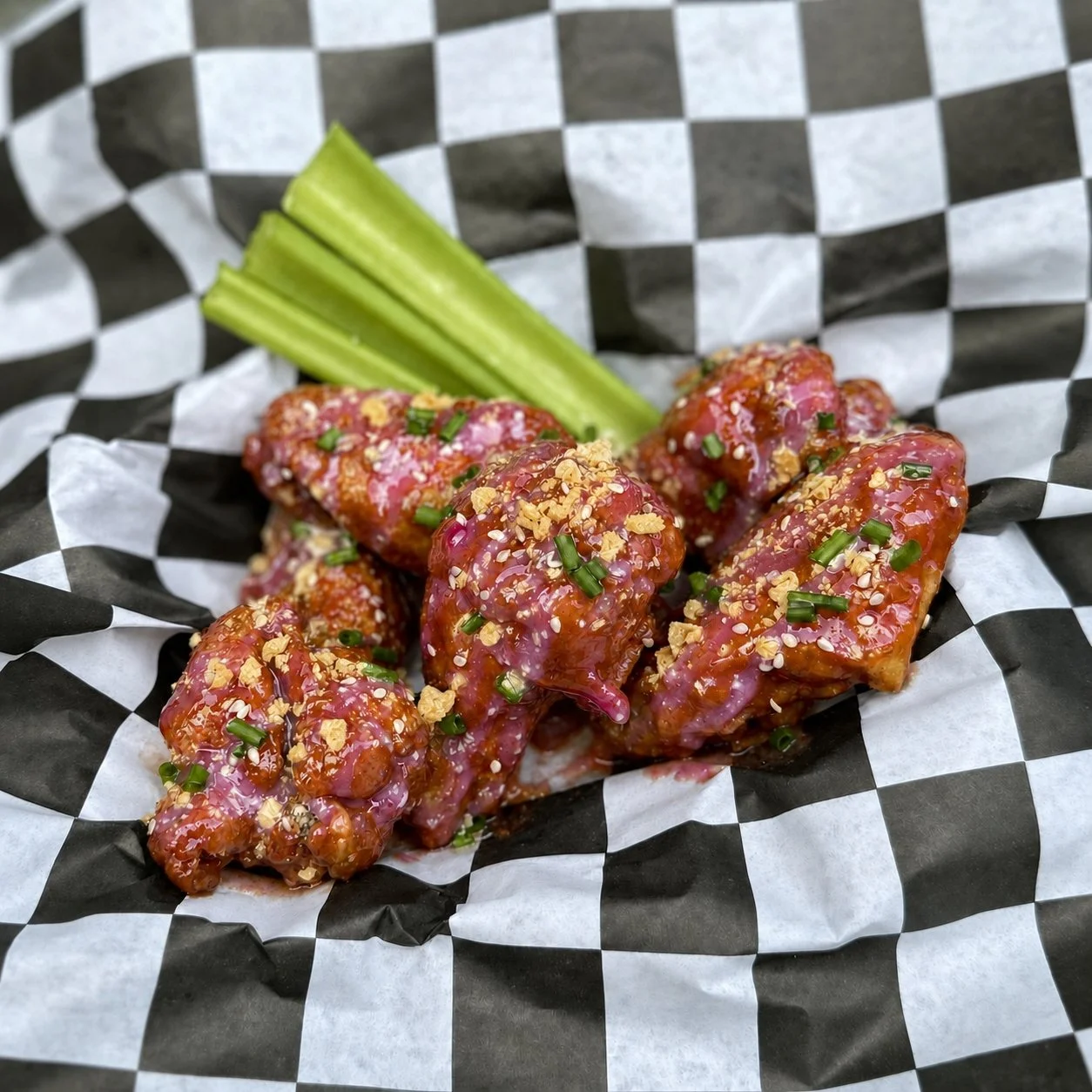 Chicken wings on top of black and white checkered liner. The wings are with a peanut butter and jelly sauce with a cream cheese jelly sauce drizzled overtop and finished with potato chips, chives and sesame seeds.
