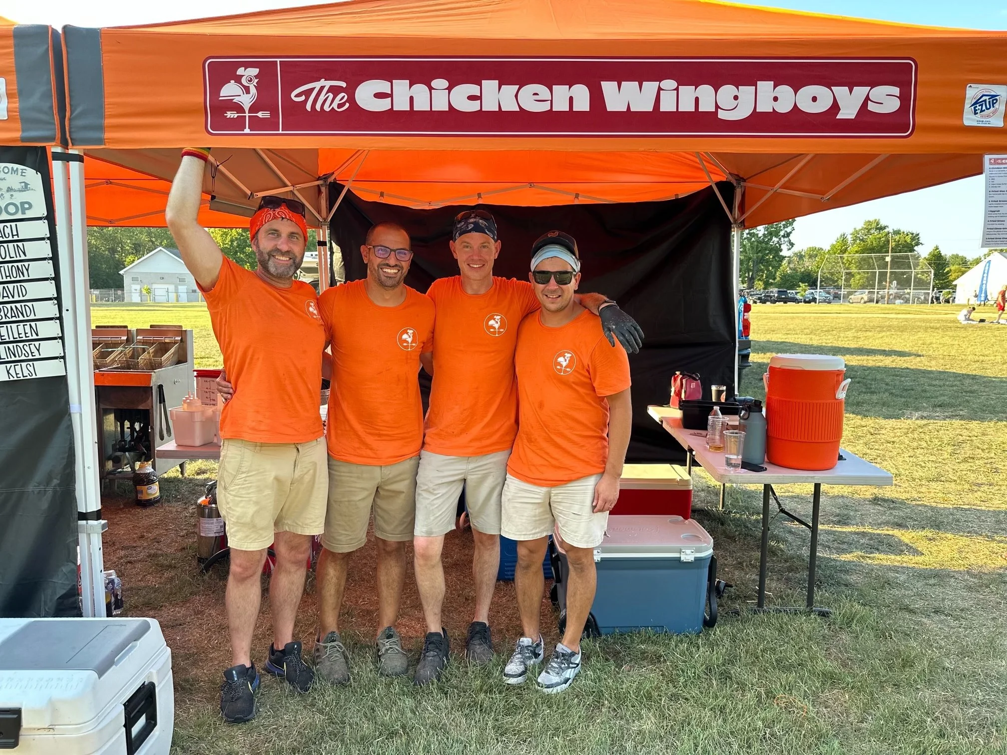 Four men standing together under an orange tent with a sign that reads 'The Chicken Wingboys'. All are wearing matching orange shirts and shorts, with sunglasses and headbands. They are smiling with their arms around each other at an outdoor event on a grassy field.