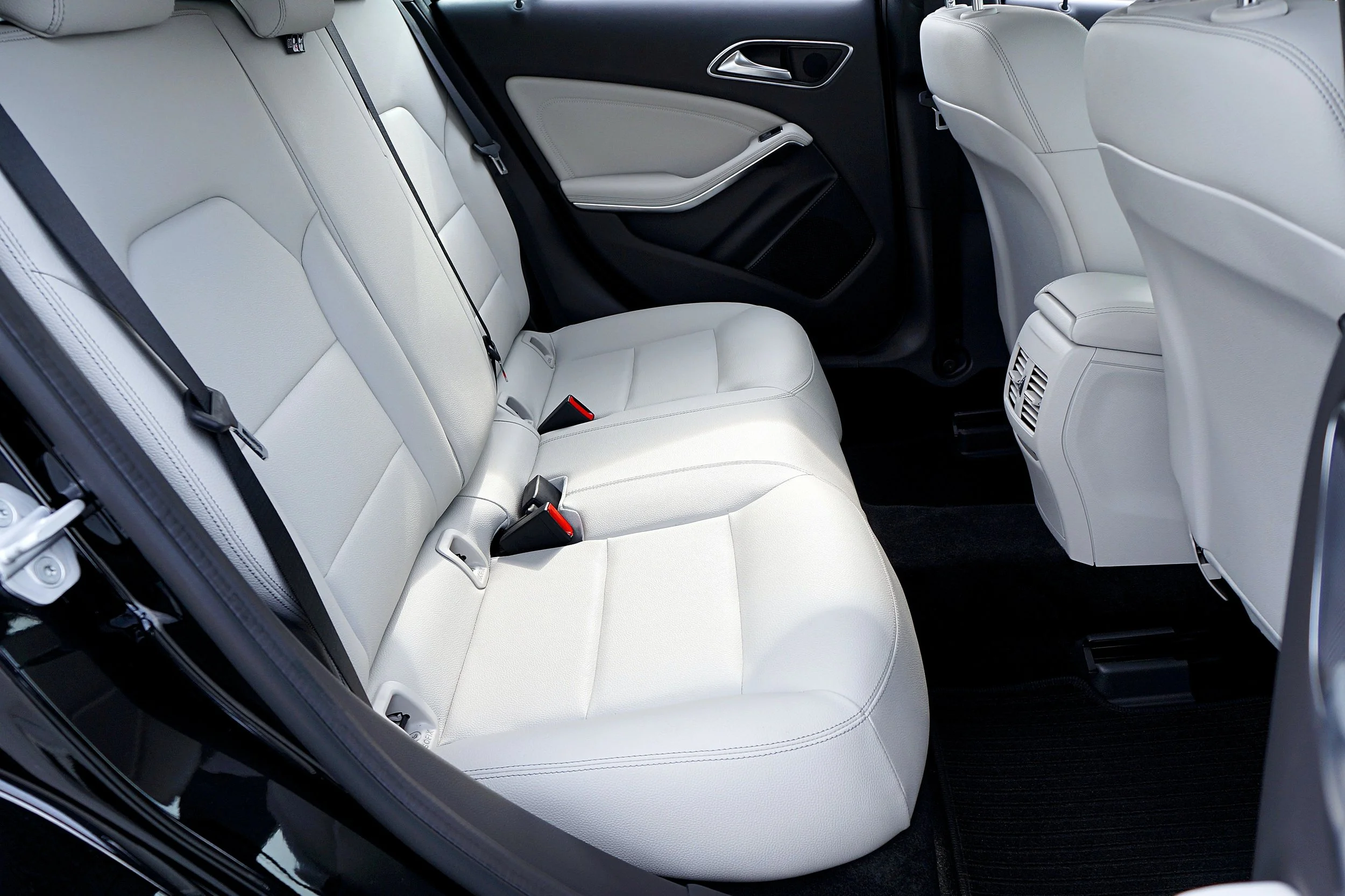 The back seat of a car with white leather upholstery, black seat belts, and a black door panel.