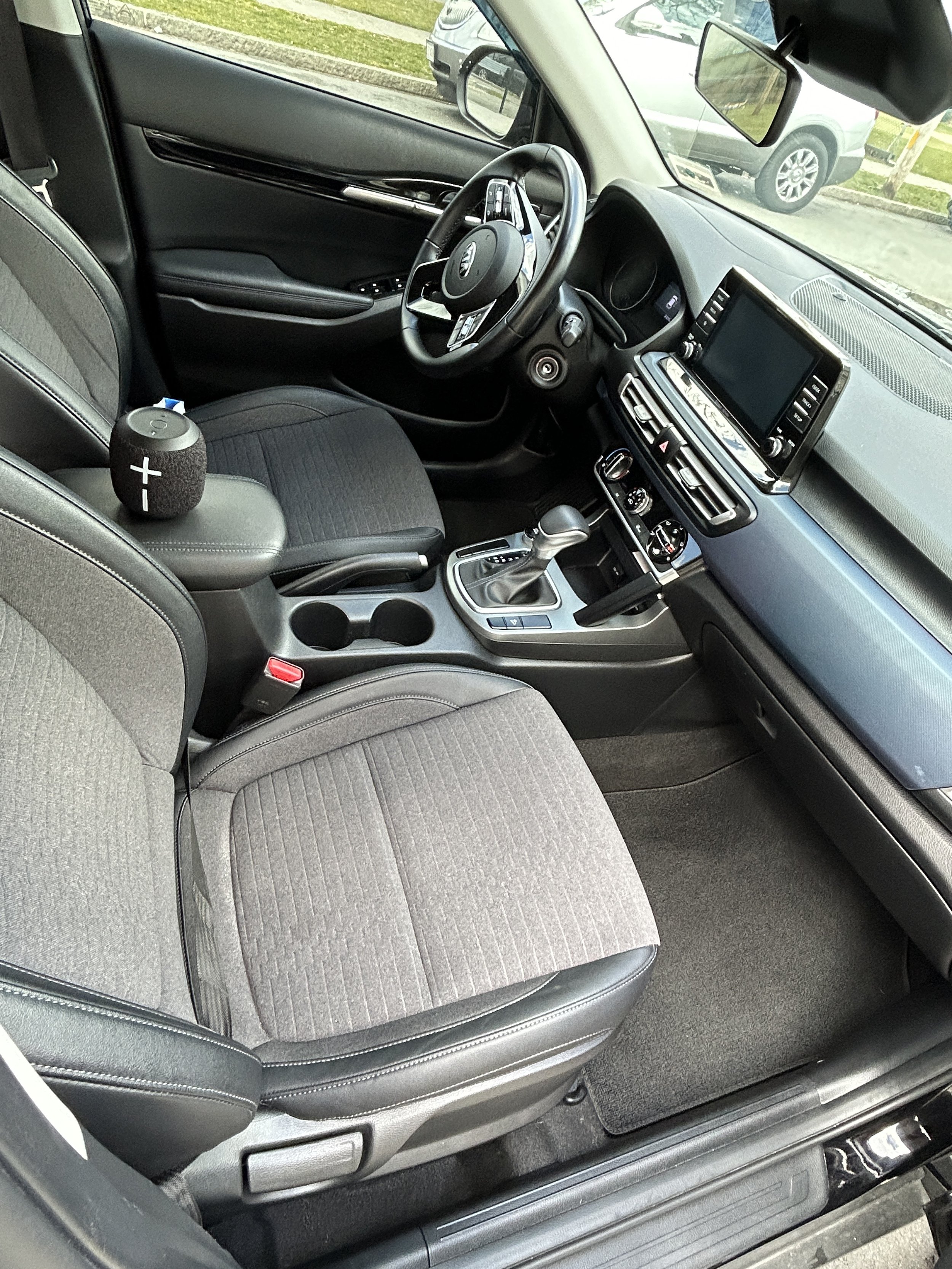 Interior of a modern vehicle with a black dashboard, steering wheel, gear shift, and gray fabric seats, with a portable speaker on the driver’s seat armrest.