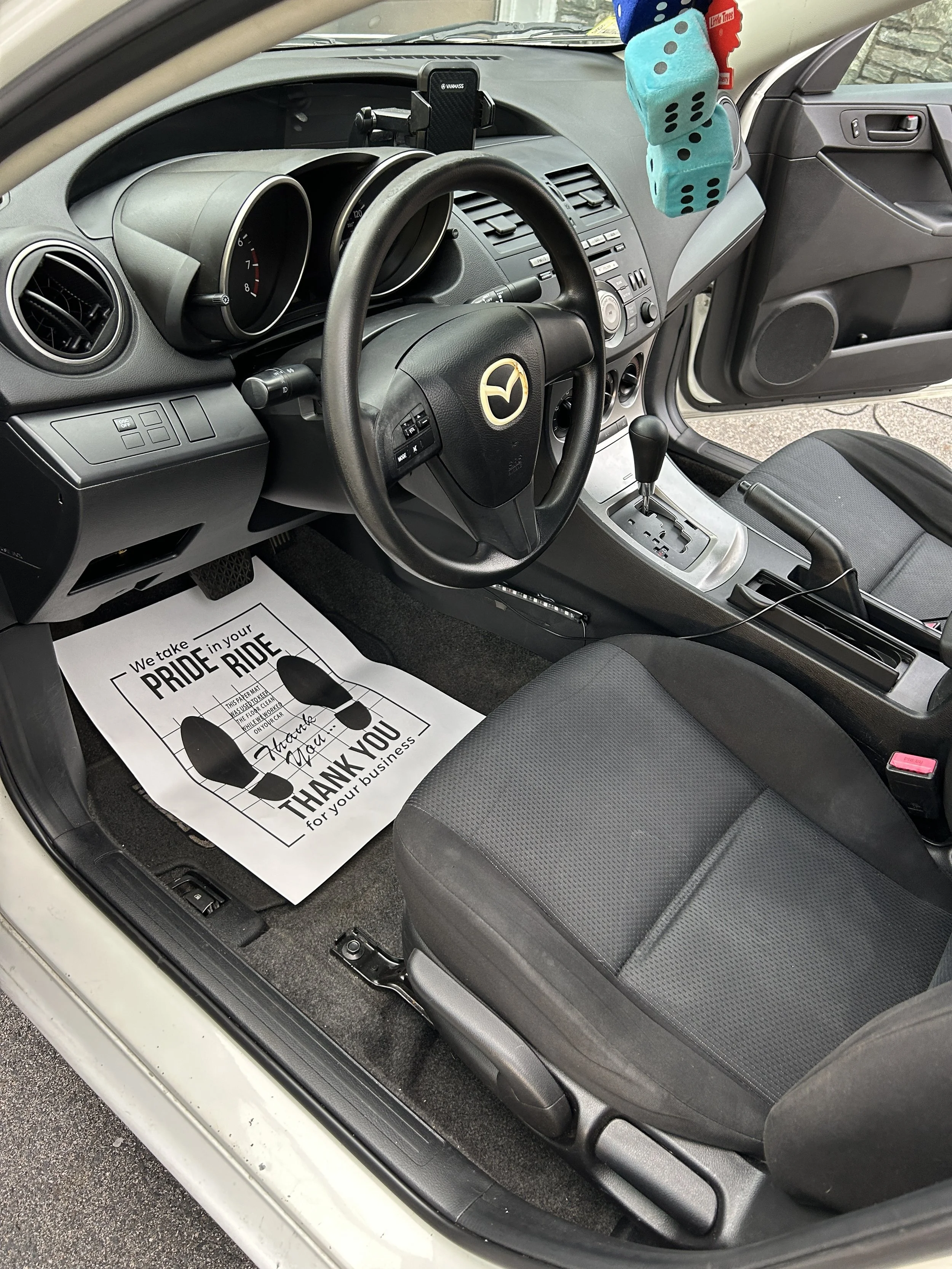 Interior of a Mazda car showing the driver's seat, steering wheel, gear shift, and hanging dice and fuzzy dice decorations from the rearview mirror. There's a paper floor mat with a message.