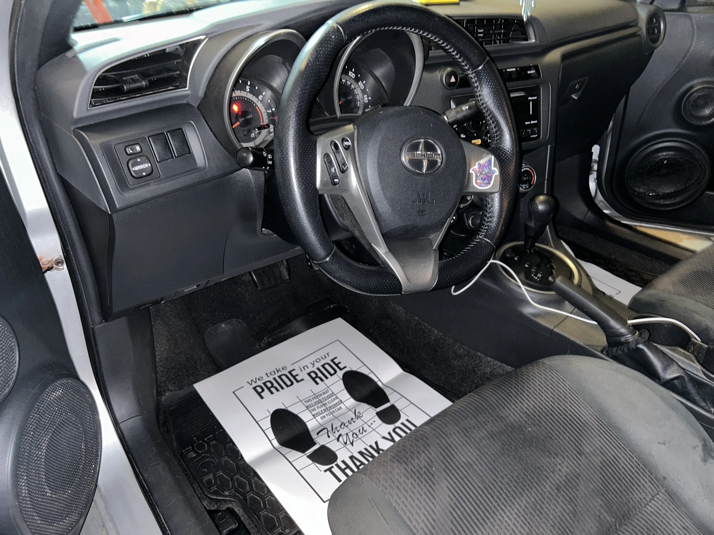 Inside a vehicle showing the dashboard, steering wheel, gear shift, driver seat, and a paper mat on the floor with a message of appreciation.