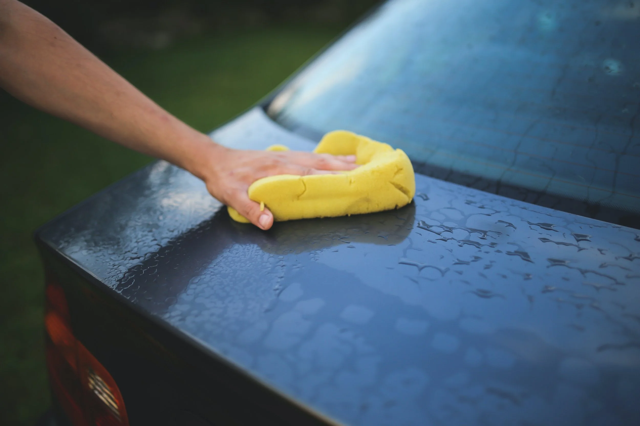 Person cleaning the hood of a car with a yellow sponge.