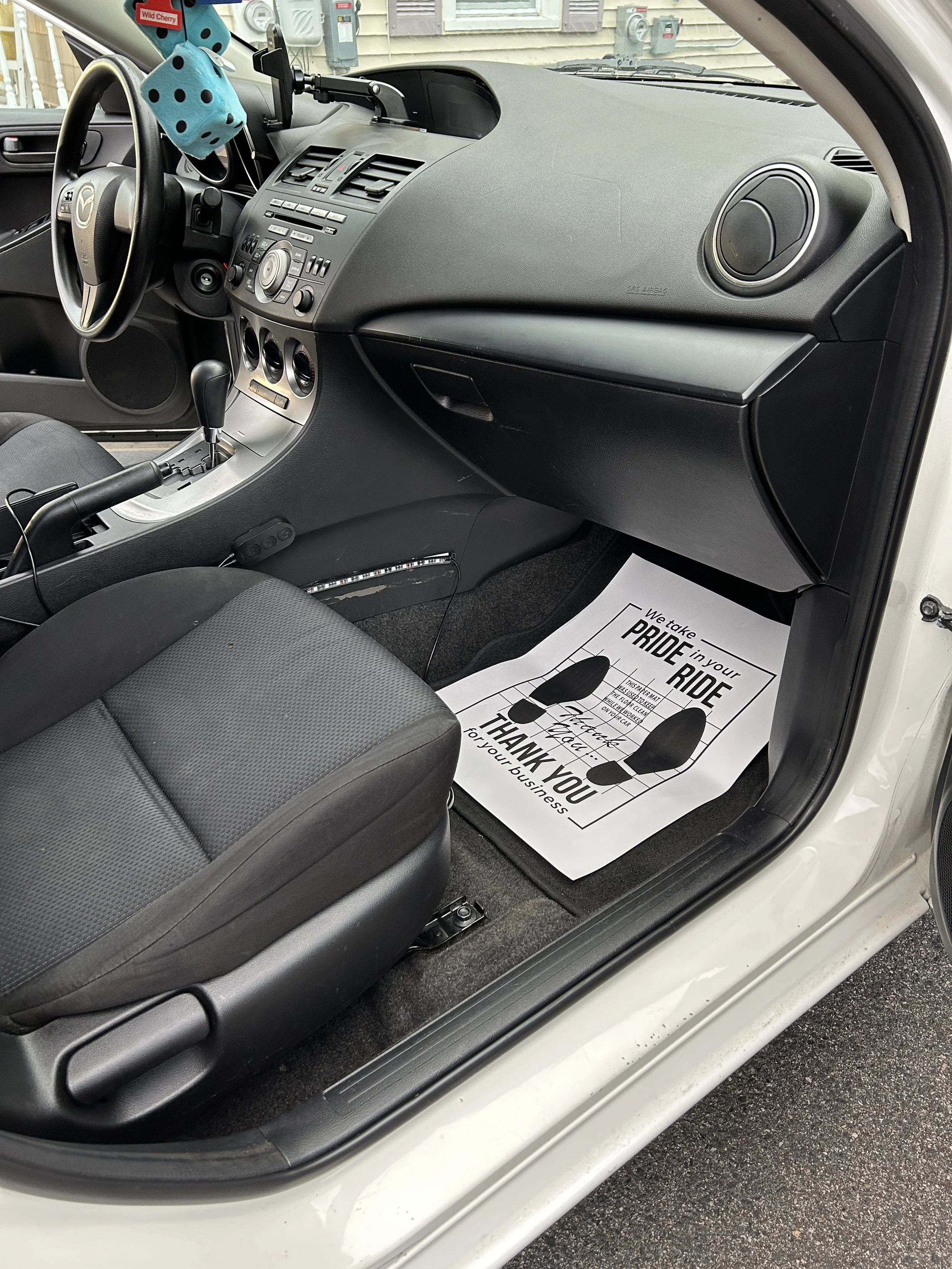 Inside a car, showing the front passenger side with a black and gray interior. There is a paper floor mat with the message "We take PRIDE in your RIDE, Thank you for your business" on the floor.