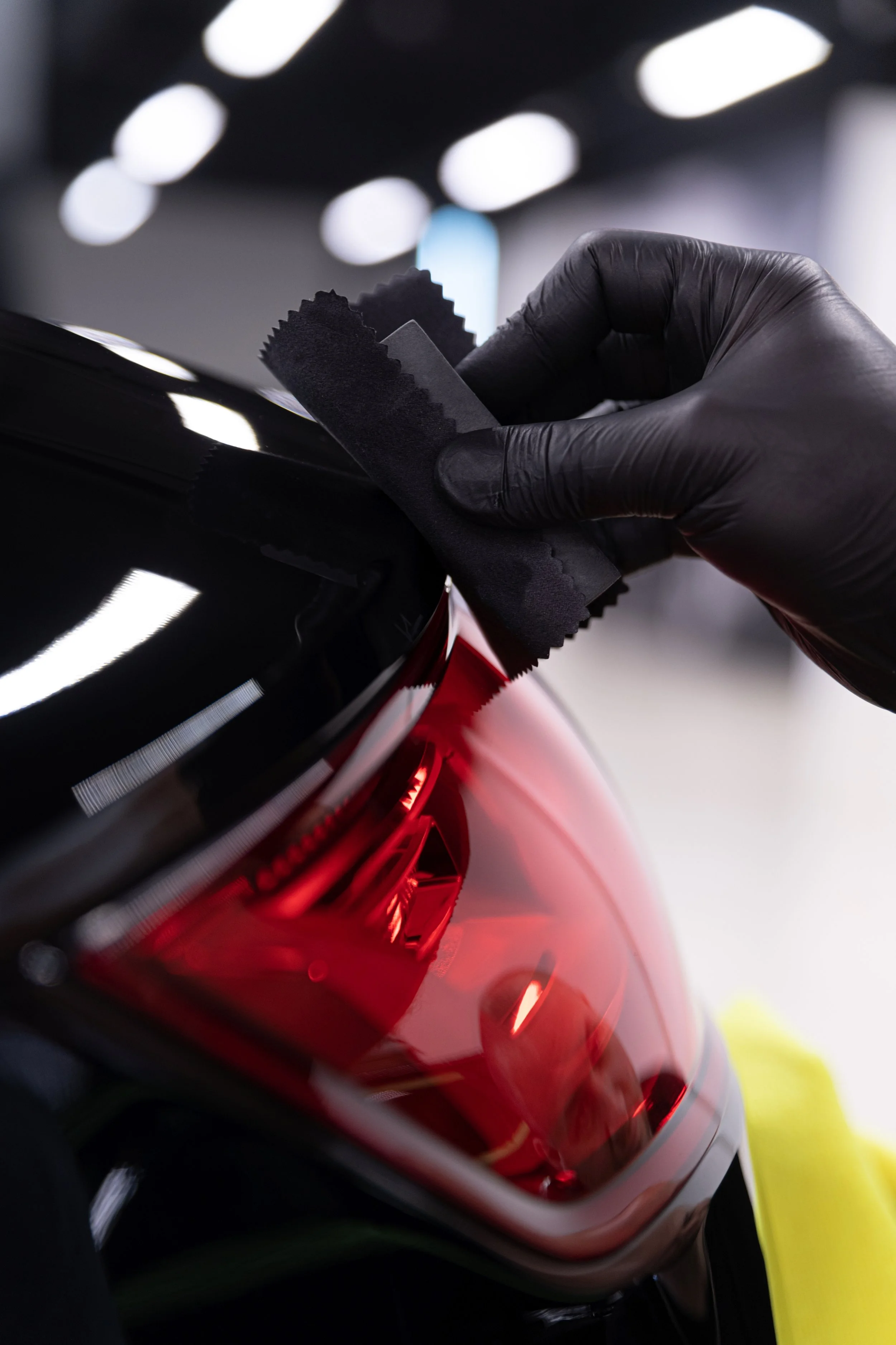 A person wearing black gloves is applying a coating or polish to a car's red taillight using a black cloth or pad.
