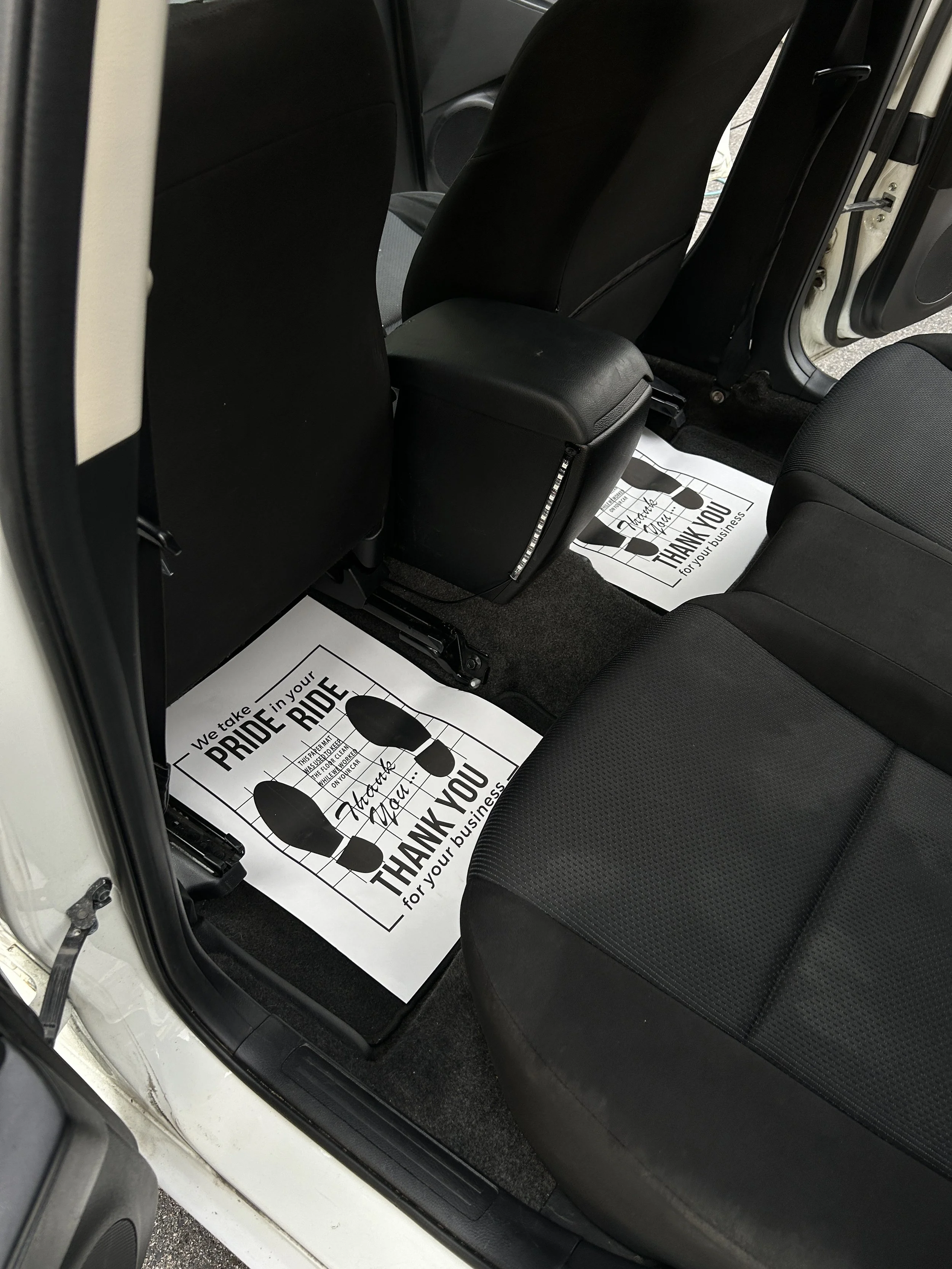 The interior of a vehicle showing black seats and floor mats with a printed message that reads, "We take pride in your ride. Thank you for your business," accompanied by an illustration of footprints.