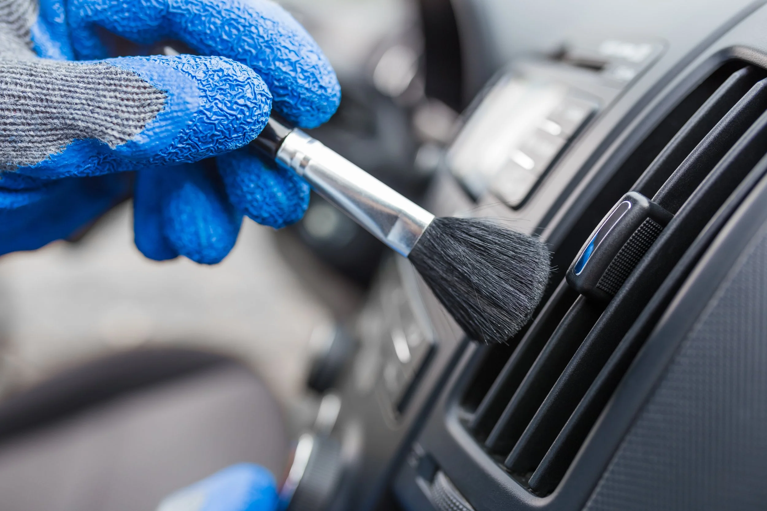A person wearing blue and gray gloves cleaning car air vents with a black brush.
