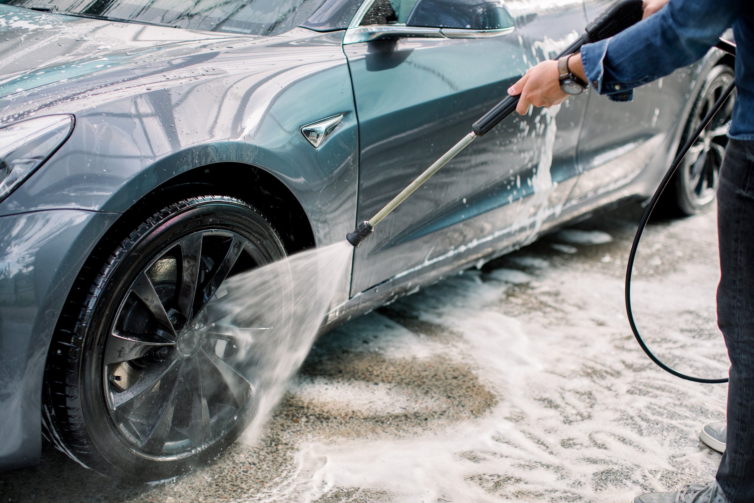 A person using a pressure washer to clean a gray Tesla car, with soap and water spraying on the vehicle.