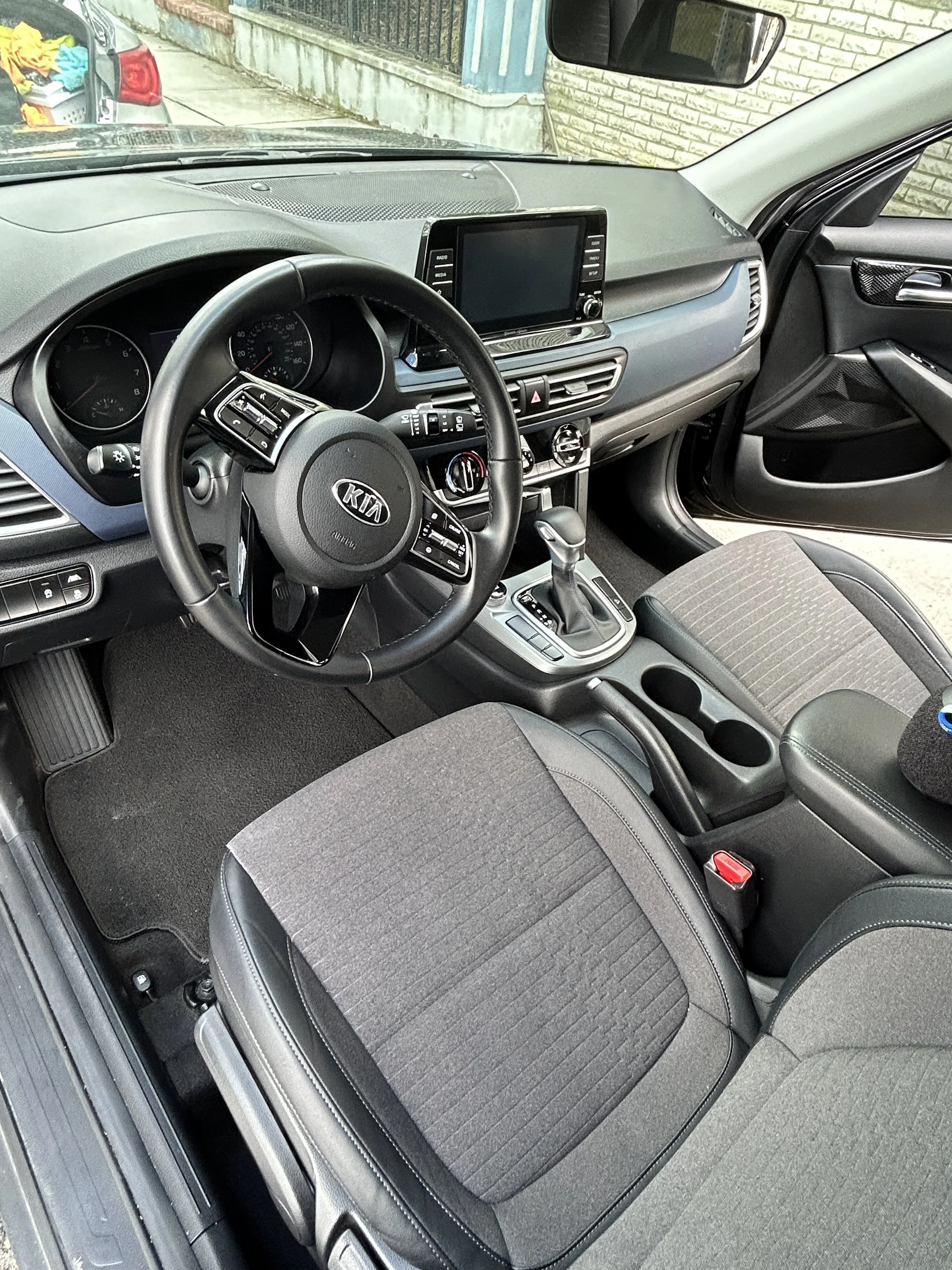 Interior of a Kia car, showing steering wheel, dashboard, touchscreen display, gear shift, and front seats.