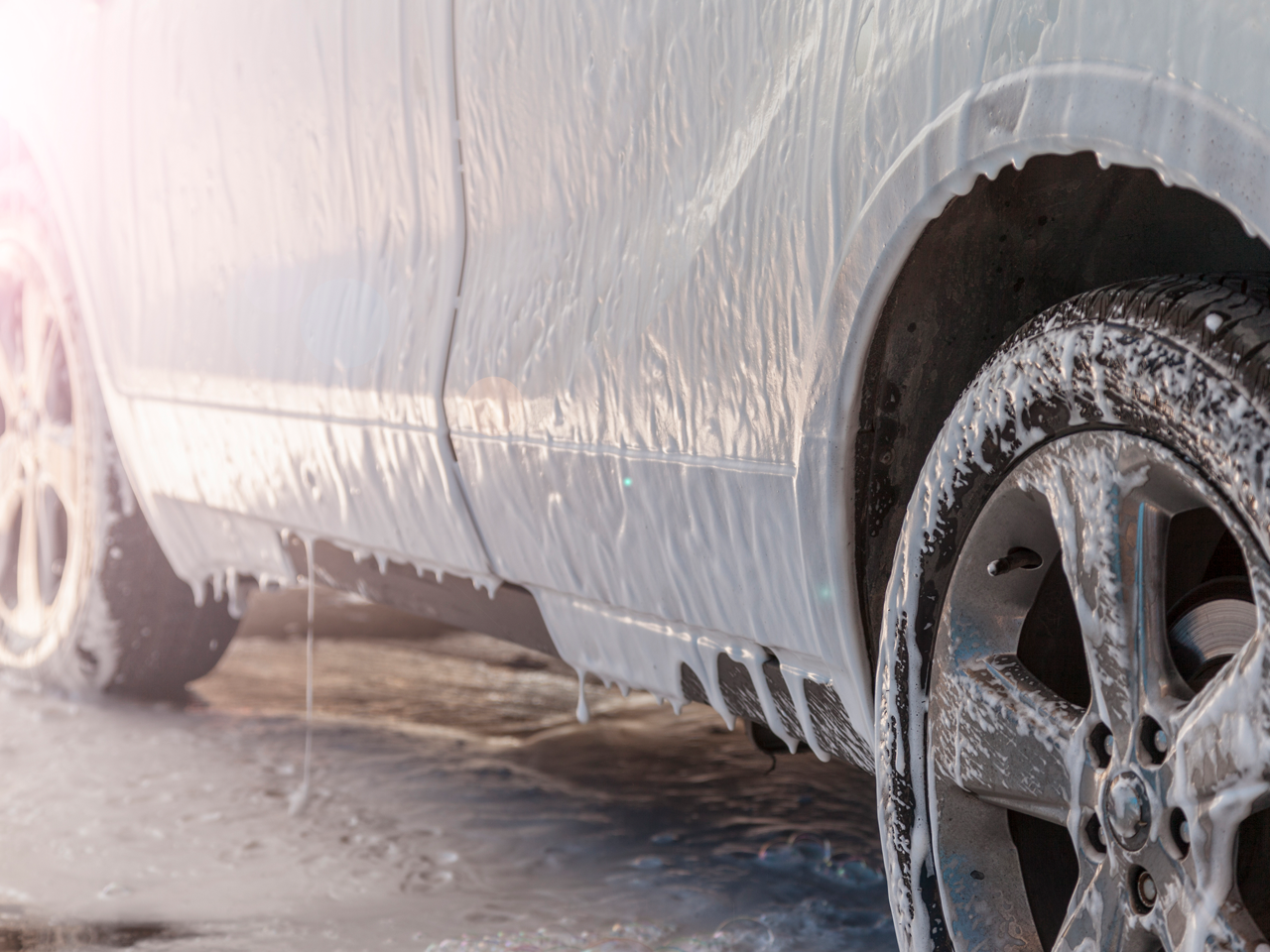 A car being washed with soap and water, covered in foam, with the wheels visible.