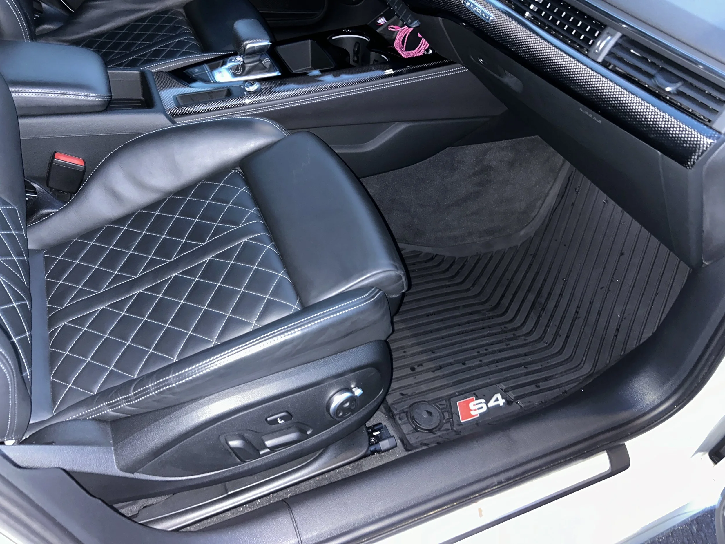 Interior of an Audi S4 with black quilted leather seat, gear shifter, dashboard, and black rubber floor mat with S4 logo.