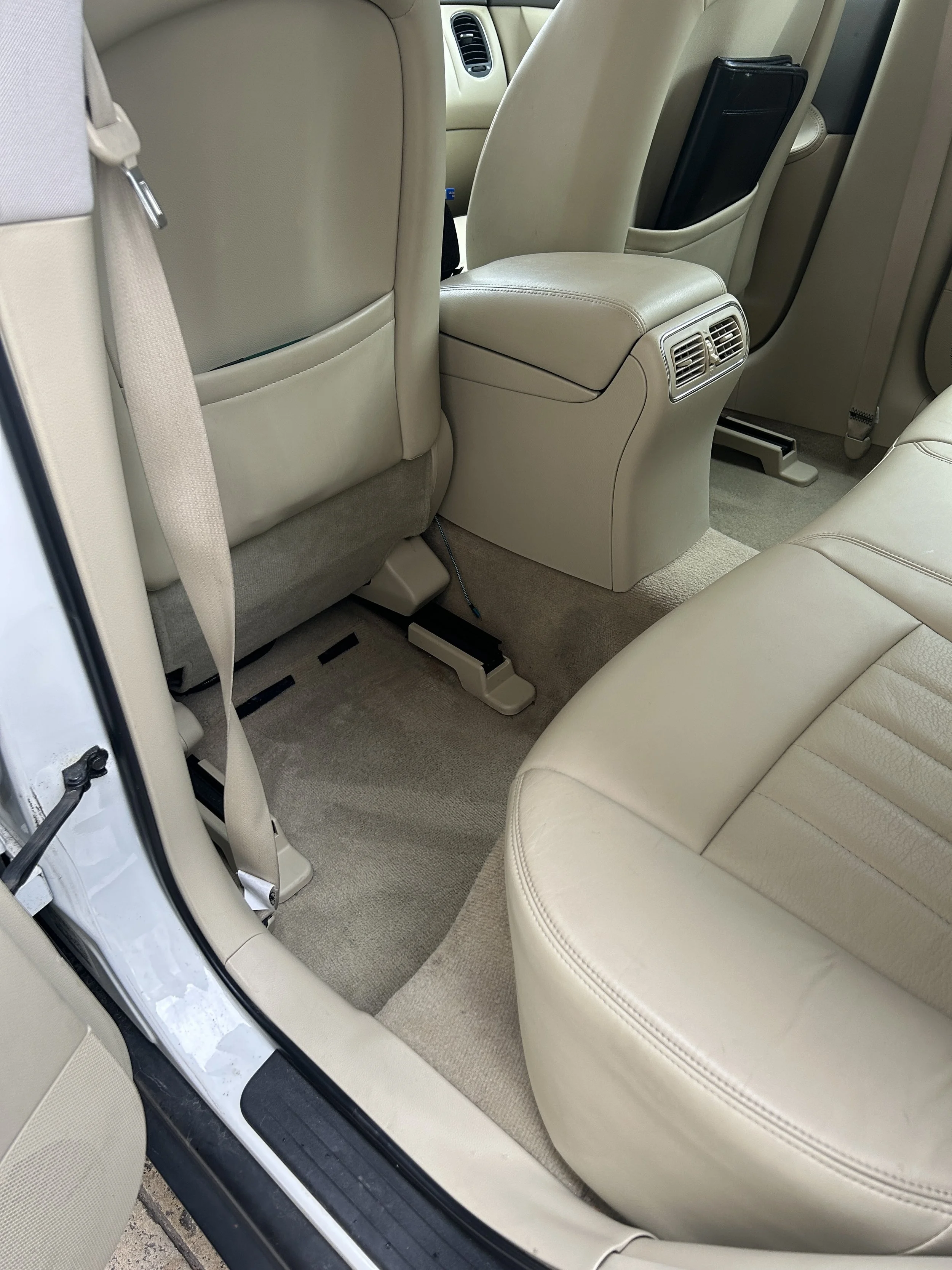 The interior of a car showing the back seat area with beige leather seats, air vents at the back of the center console, and beige carpeting.