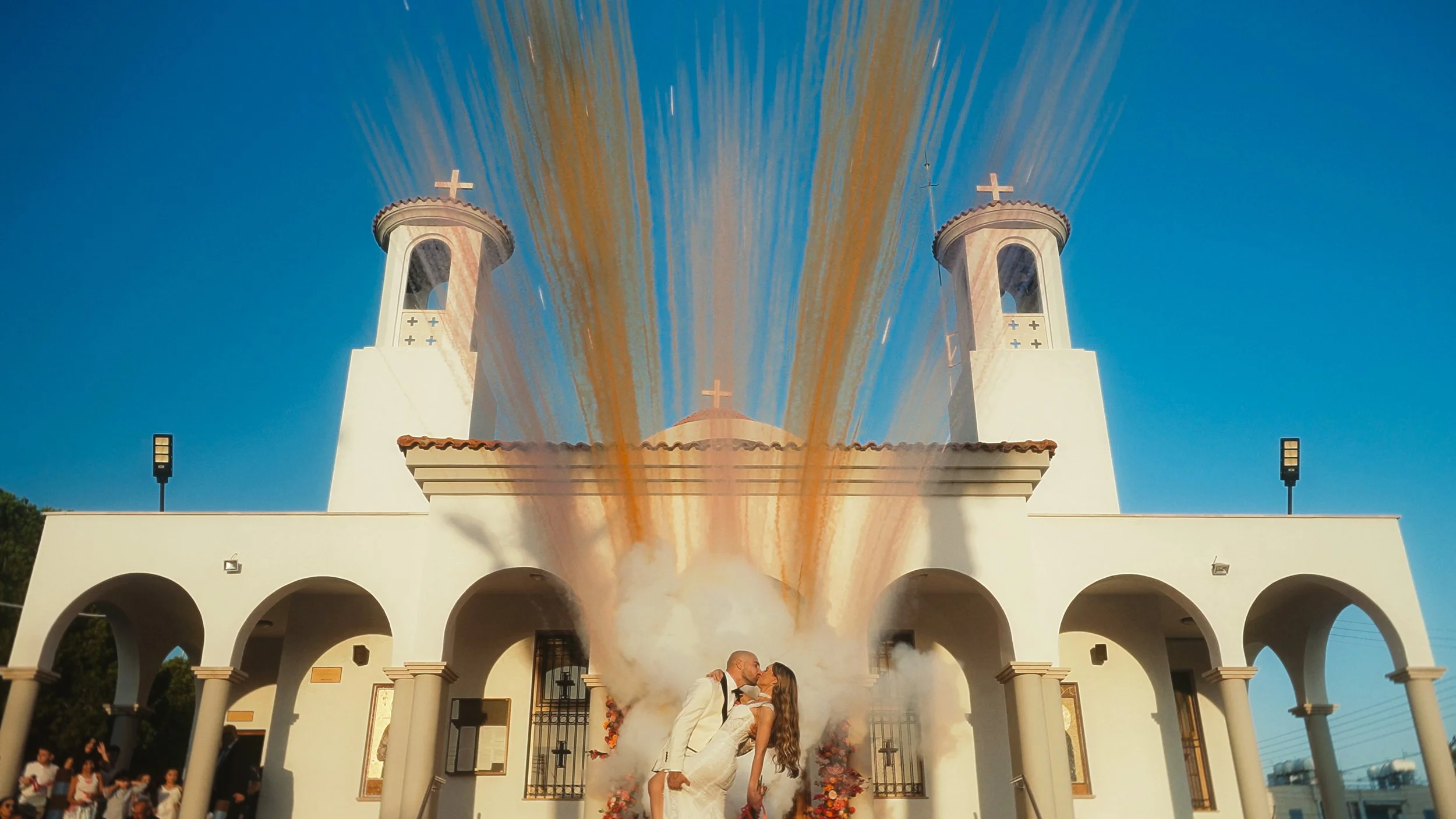 Couple kissing outside church with colourful daytime fireworks against blue sky, Ayios Stylianos Church Agios Athanasios Cyprus, Red Lens Films