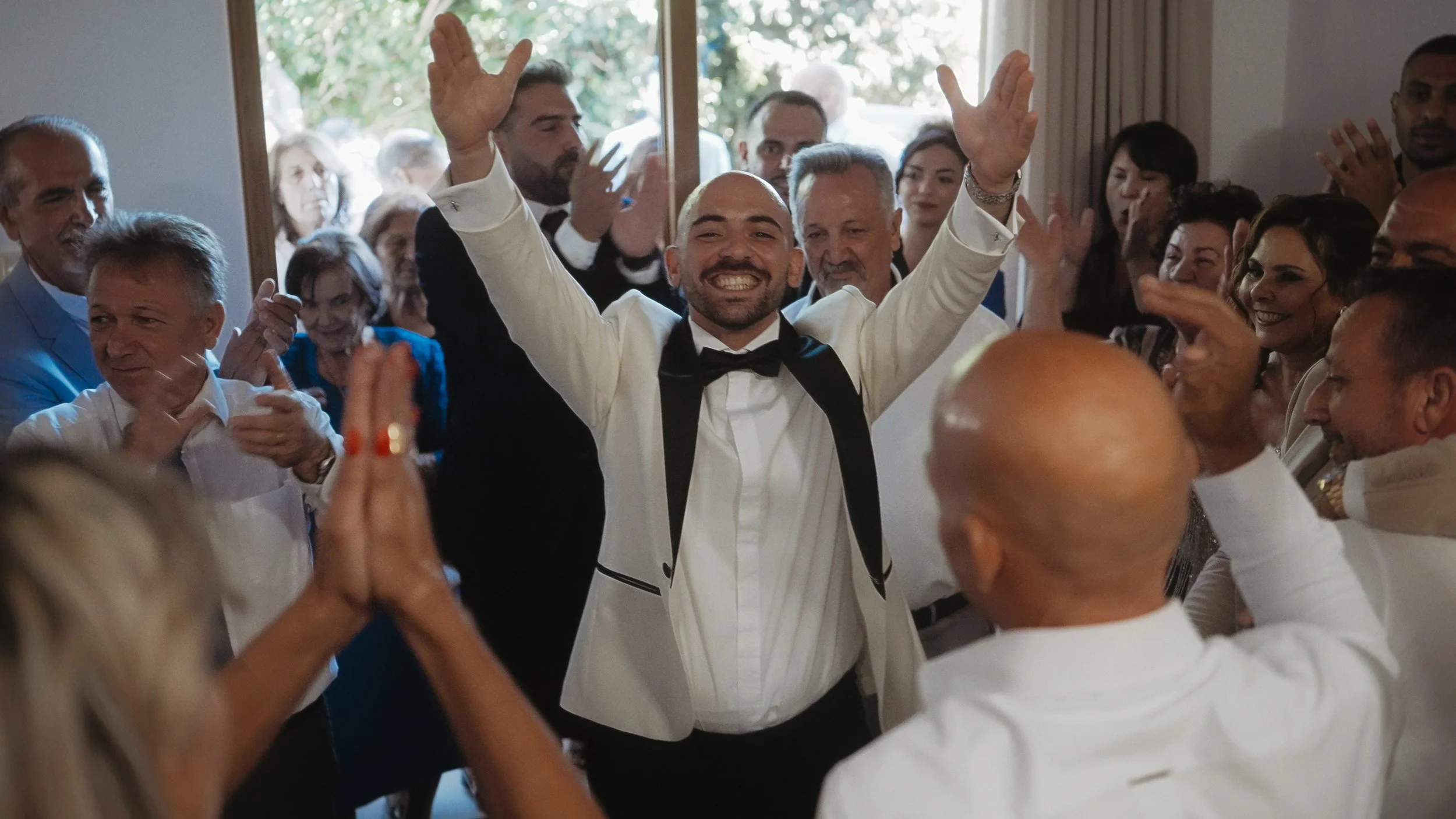 Groom celebrating with arms raised surrounded by cheering guests during traditional Cypriot preparation, Kato Polemidia Cyprus, Red Lens Films