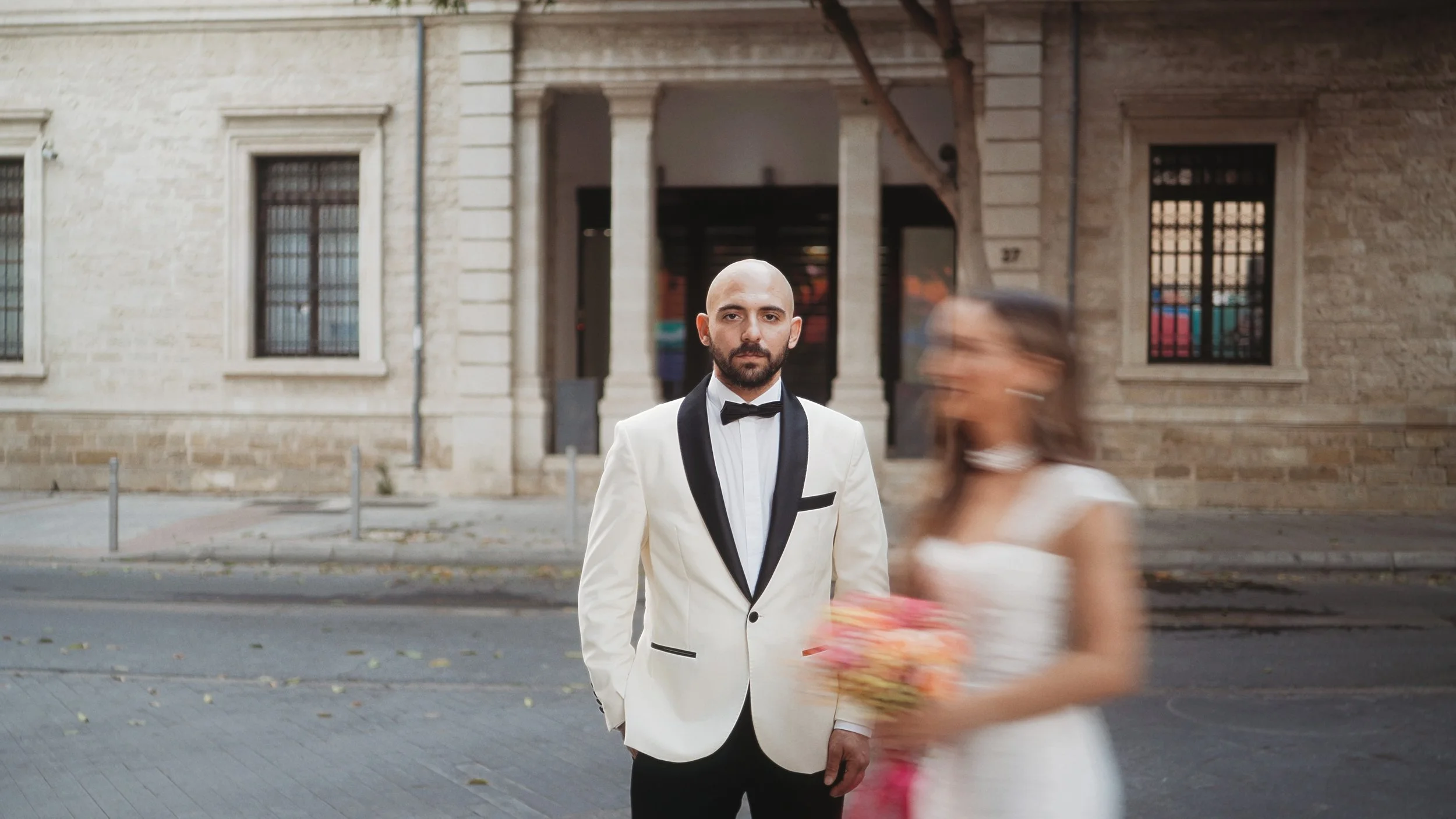 Groom in white tuxedo standing still while bride walks past with bouquet, couple portrait in old town Limassol, Red Lens Films