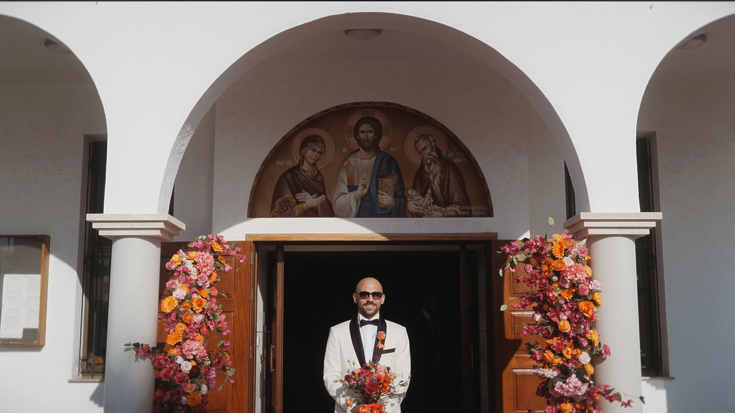 Groom at church entrance with fuchsia and orange flower arrangements and Byzantine mosaic, Ayios Stylianos Church Agios Athanasios Cyprus, Red Lens Films