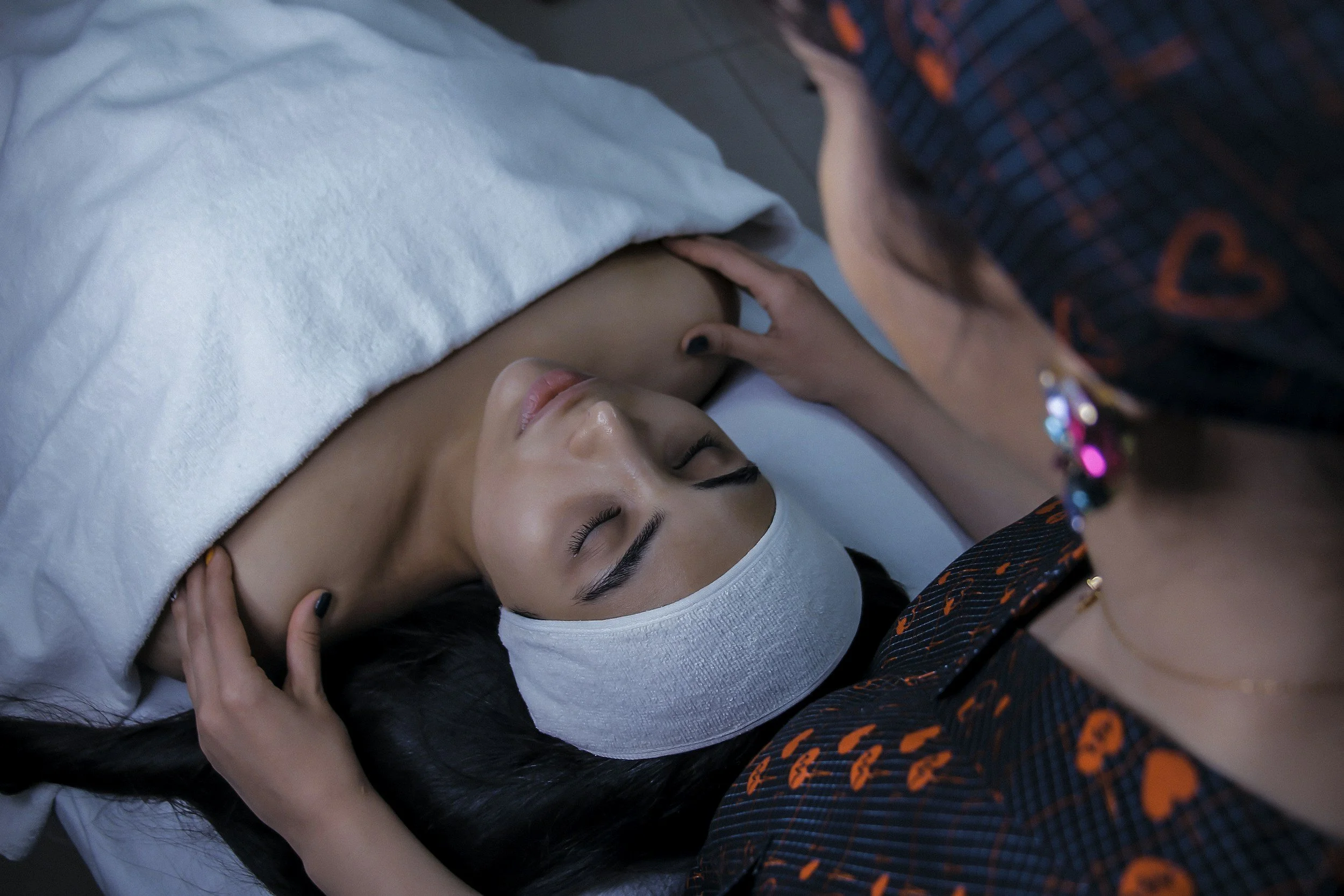 A woman lying down with a white headband on her forehead, eyes closed, receiving a massage or treatment on her neck and shoulder area from a practitioner, who is wearing a dark shirt with orange patterns and a colorful beaded necklace.