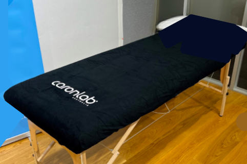 A massage or treatment table with a black cover, a pillow at the head, and wooden legs, situated on a wooden floor in a room.