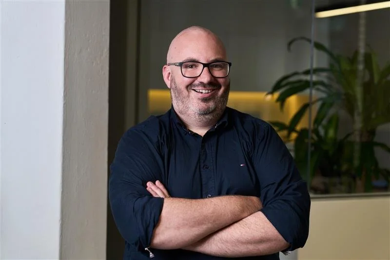 Man in black t-shirt leaning against tree in outdoor setting.